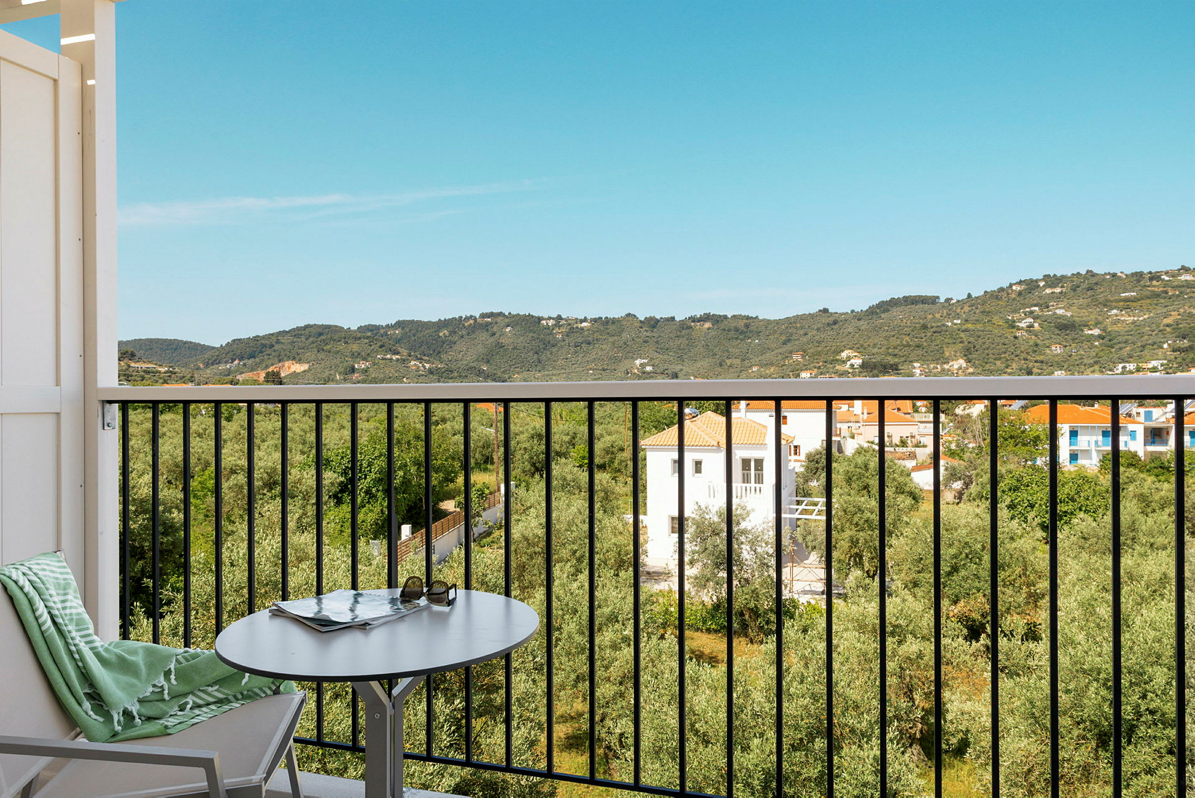 Udsigt fra en balkon med bord og stol over et frodigt landskab med huse og bjerge. (AI-genereret tekst)