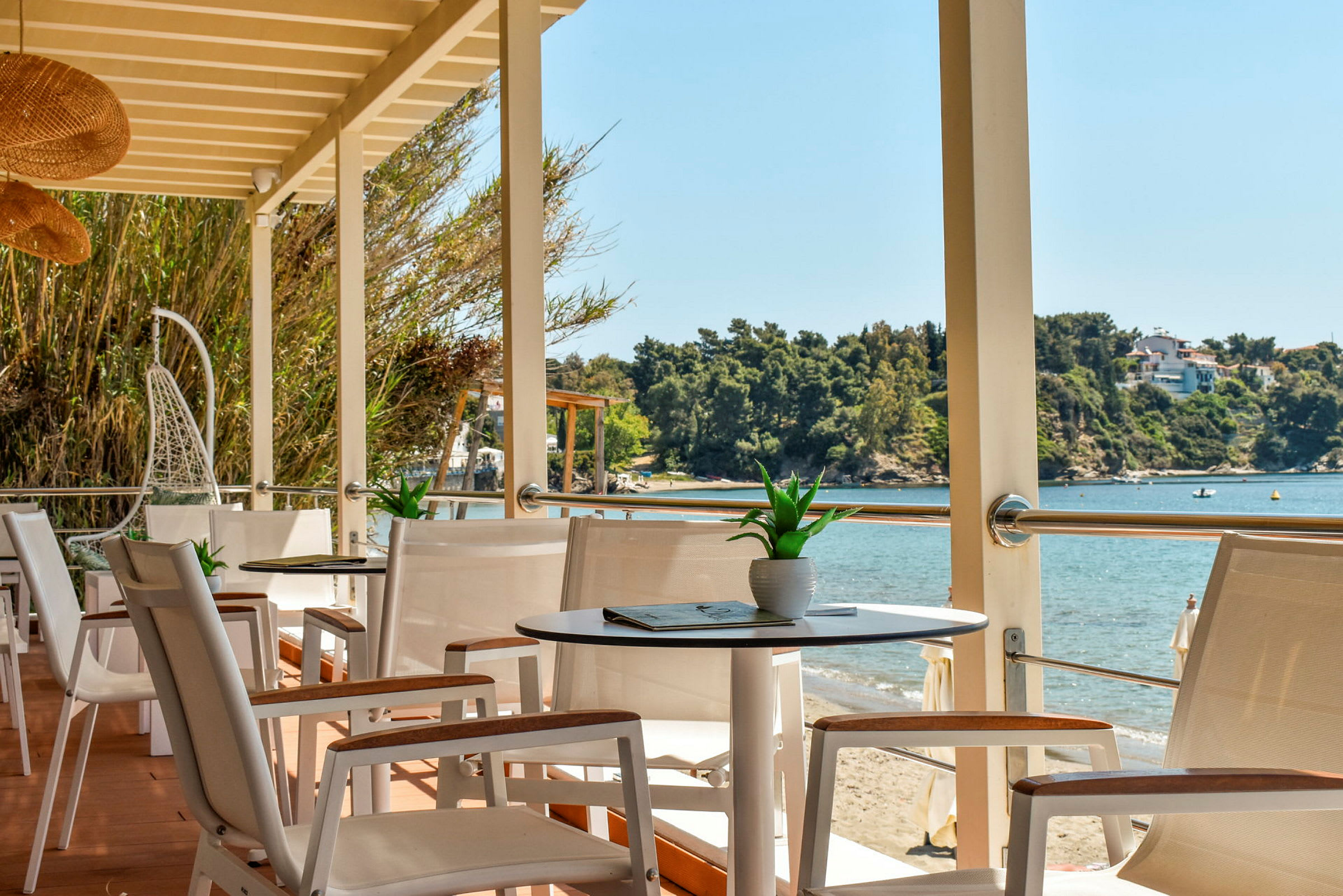 Udsigt fra en terrasse med borde og stole mod en strand og havet. (AI-genereret tekst)
