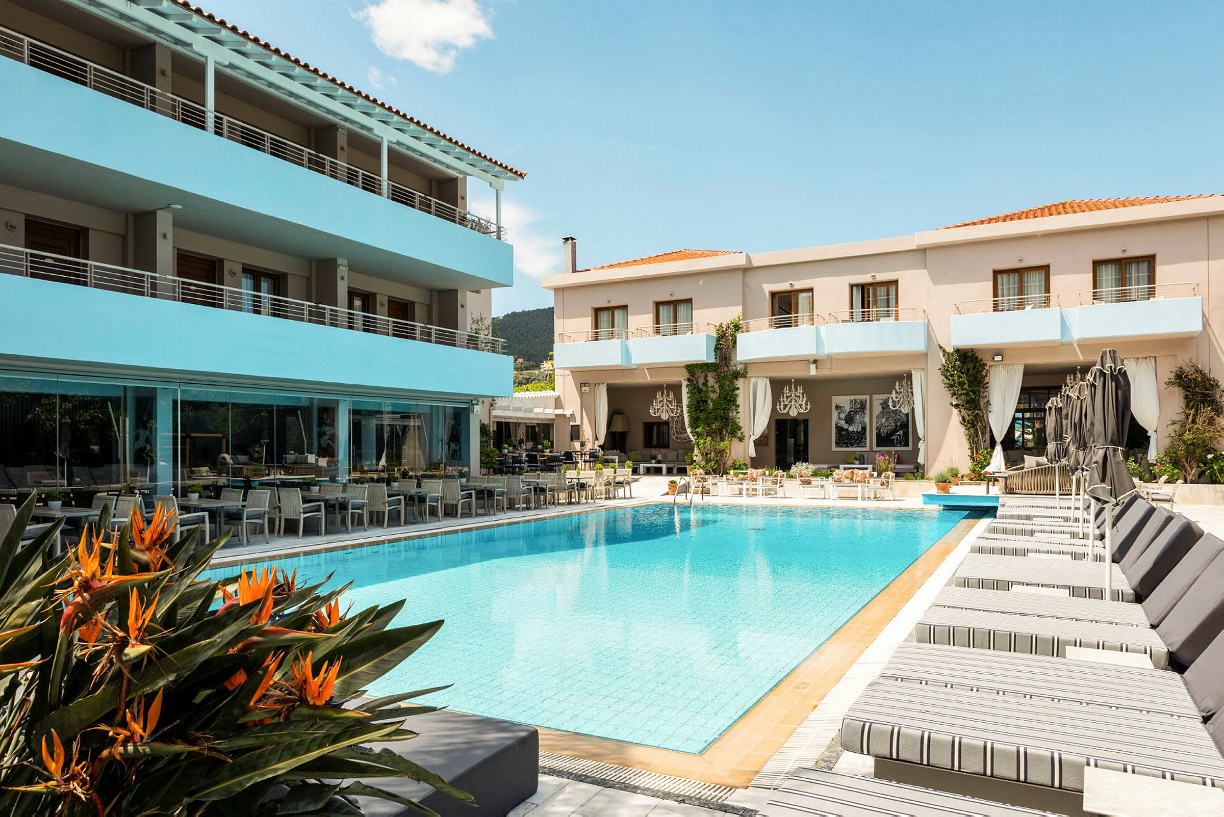 Et hotel med en pool i forgrunden, omgivet af solsenge og nogle orange blomster tættest på billedet. Bygningen har altaner og en udendørs serveringsplads. (AI-genereret tekst)