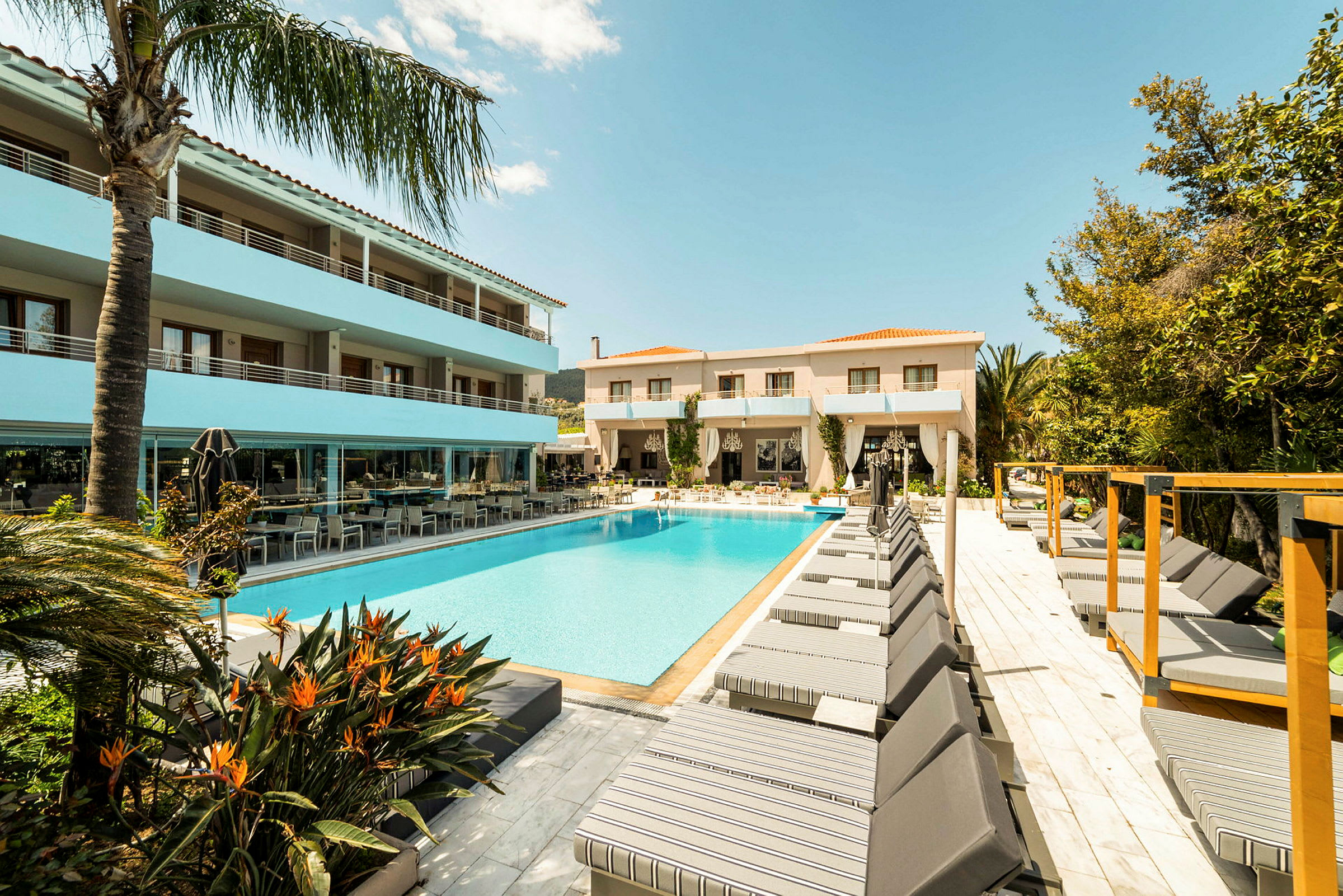 En hotelpool omgivet af liggestole og bygninger under en blå himmel. (AI-genereret tekst)