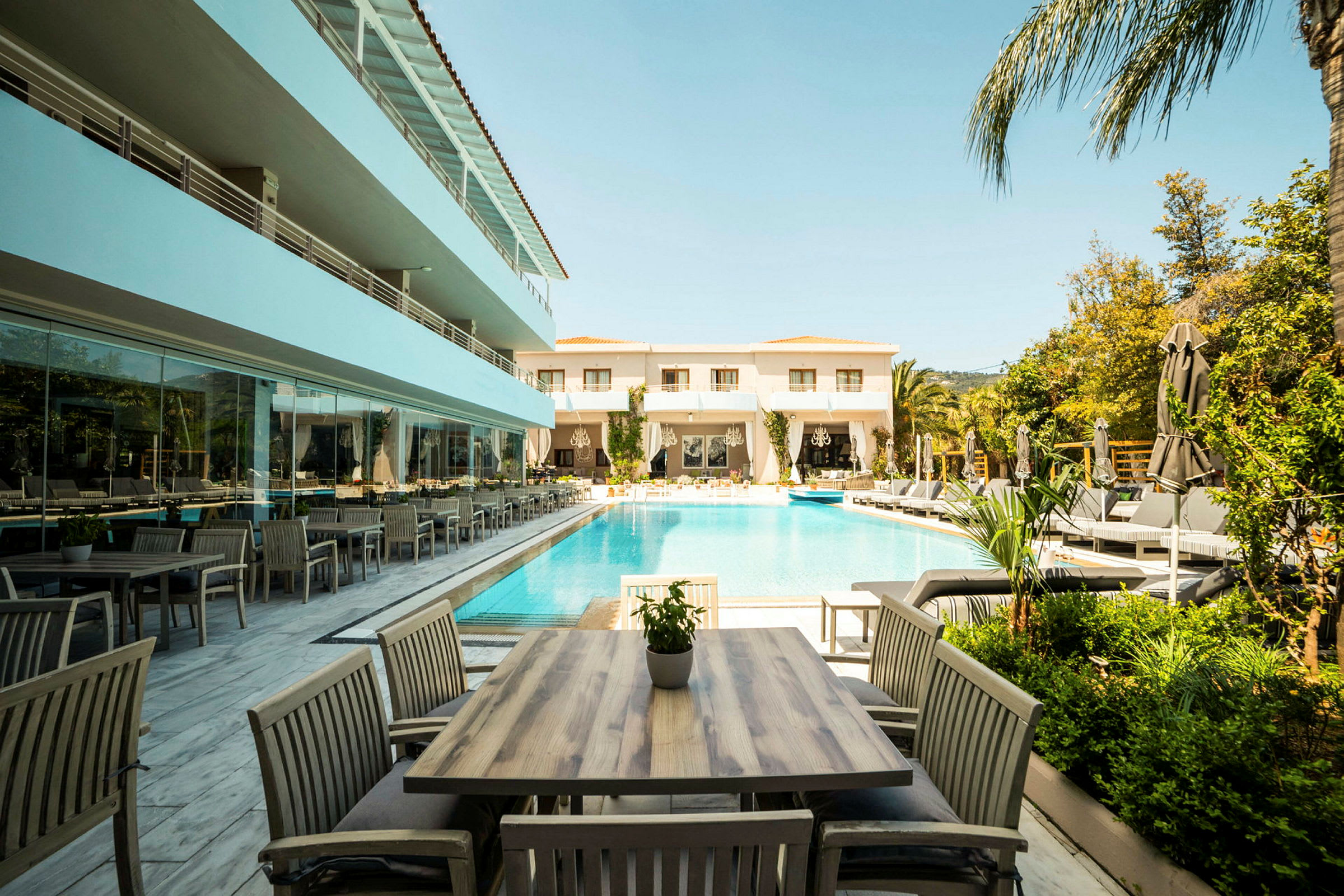 Et hotelområde med en udendørs pool omgivet af solsenge og parasoller. Borde og stole står på en terrasse ved siden af poolen. (AI-genereret tekst)