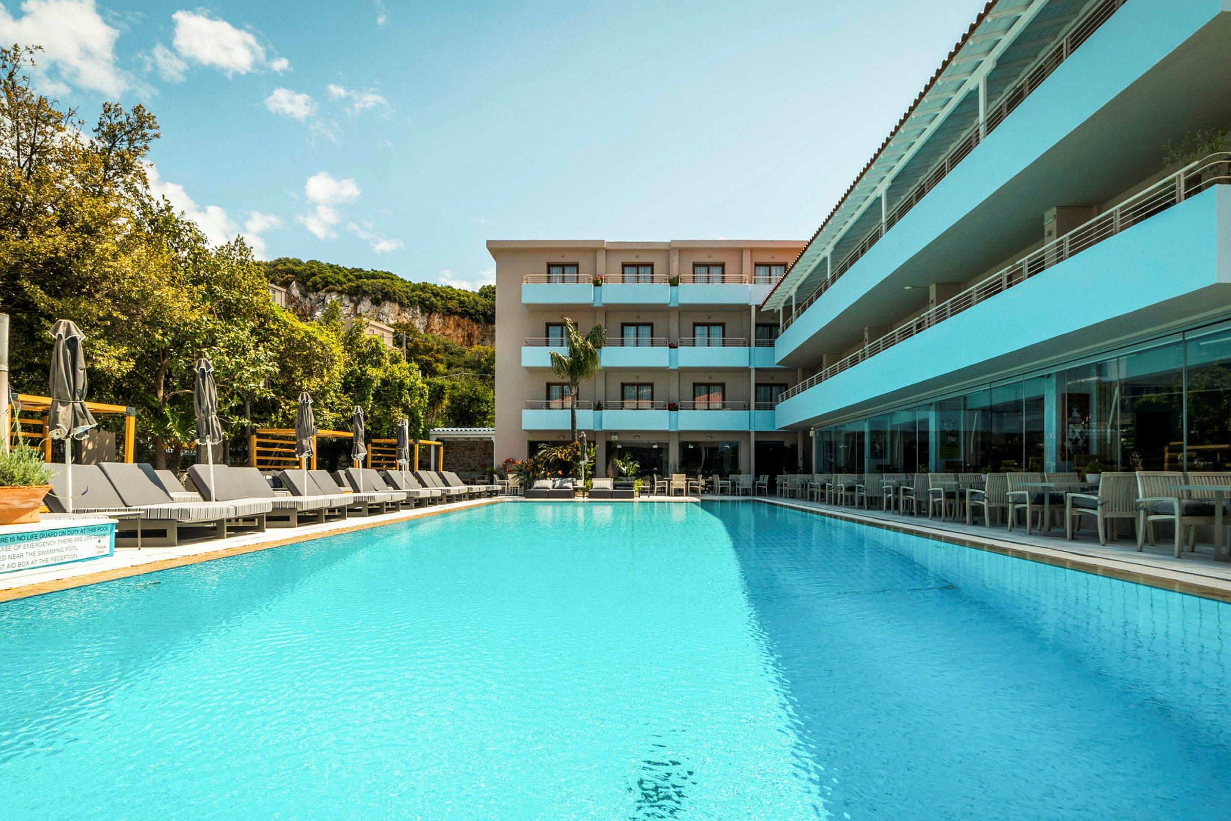 En udendørs pool foran et hotel omgivet af vegetation og klipper. (AI-genereret tekst)