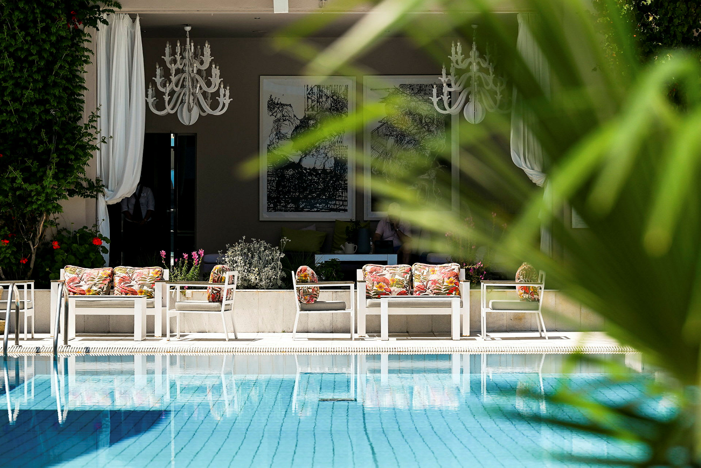 Et udendørsmiljø med en pool og siddeområde med farverige puder i forgrunden, omgivet af grønt. (AI-genereret tekst)