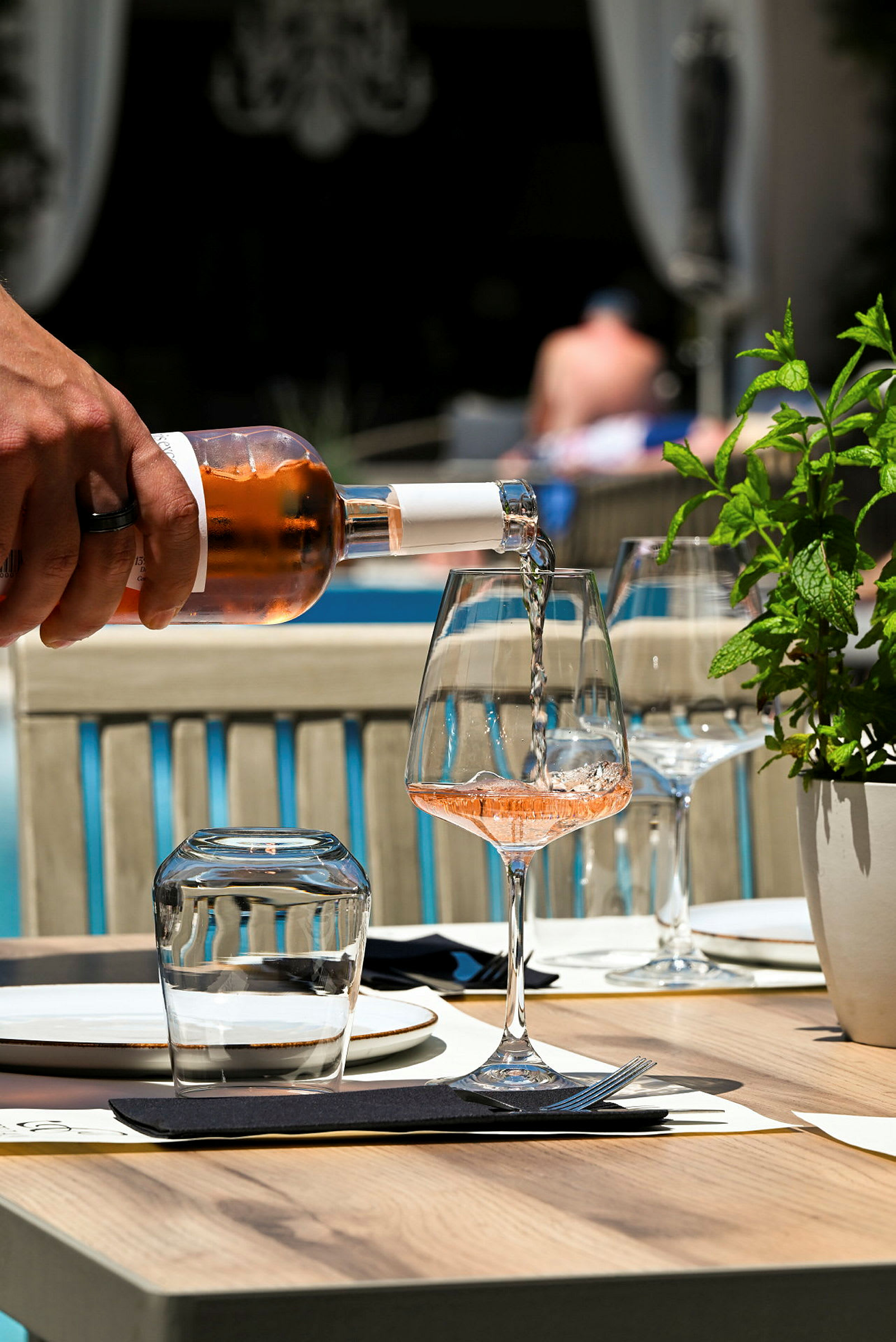 Et vinglas fyldes med rosévin ved et dækket bord ved siden af en grøn plante. (AI-genereret tekst)