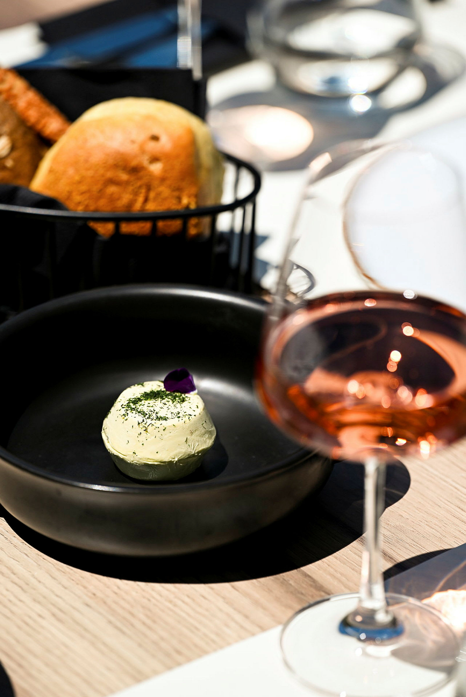 En sort tallerken med smør pyntet med en blomst, et brød i en kurv og et glas rosévin på et bord. (AI-genereret tekst)