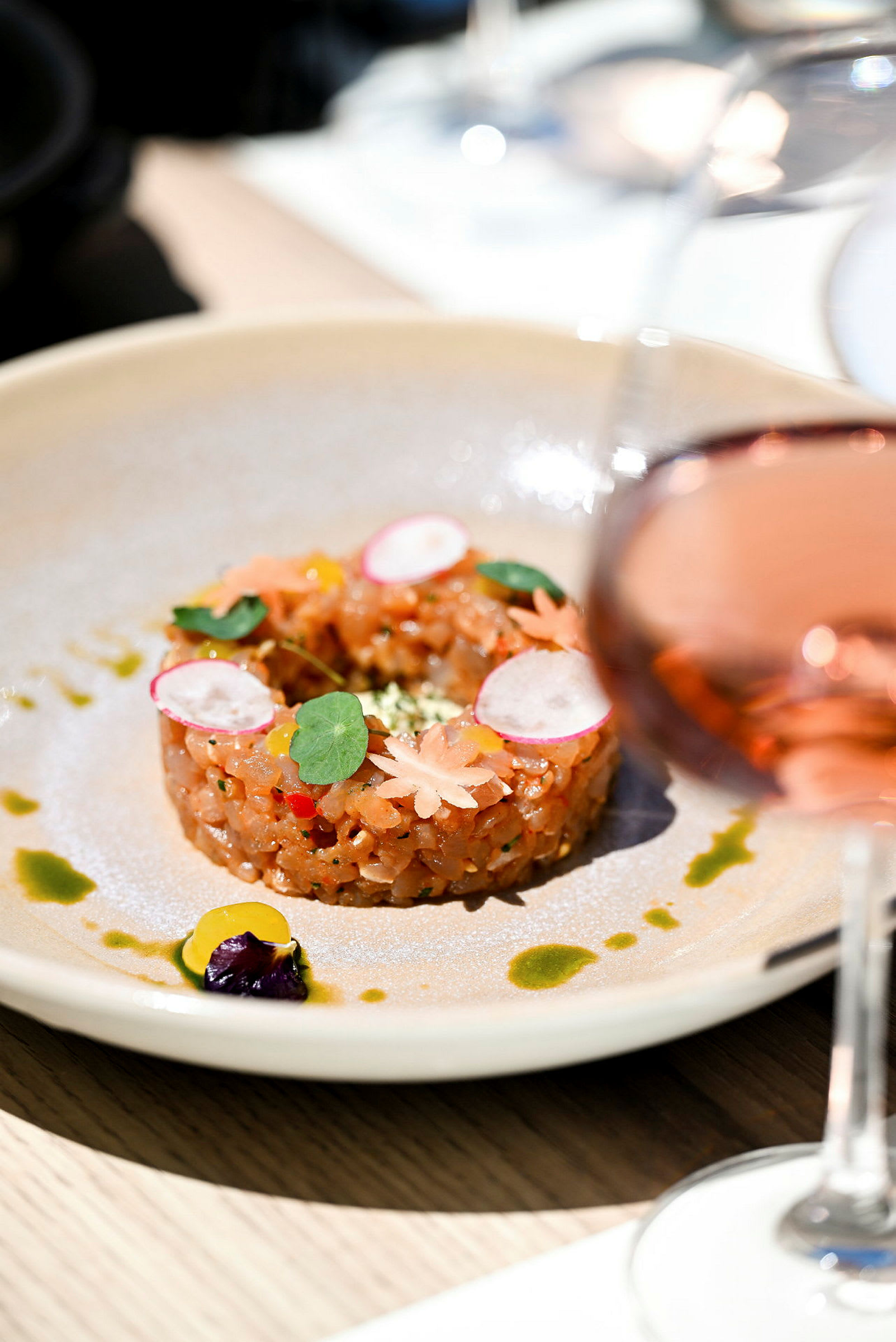 En tallerken med en dekoreret ret, der ligner en tartar, omgivet af små saucedetaljer. Et glas med en roséagtig drik i forgrunden. (AI-genereret tekst)