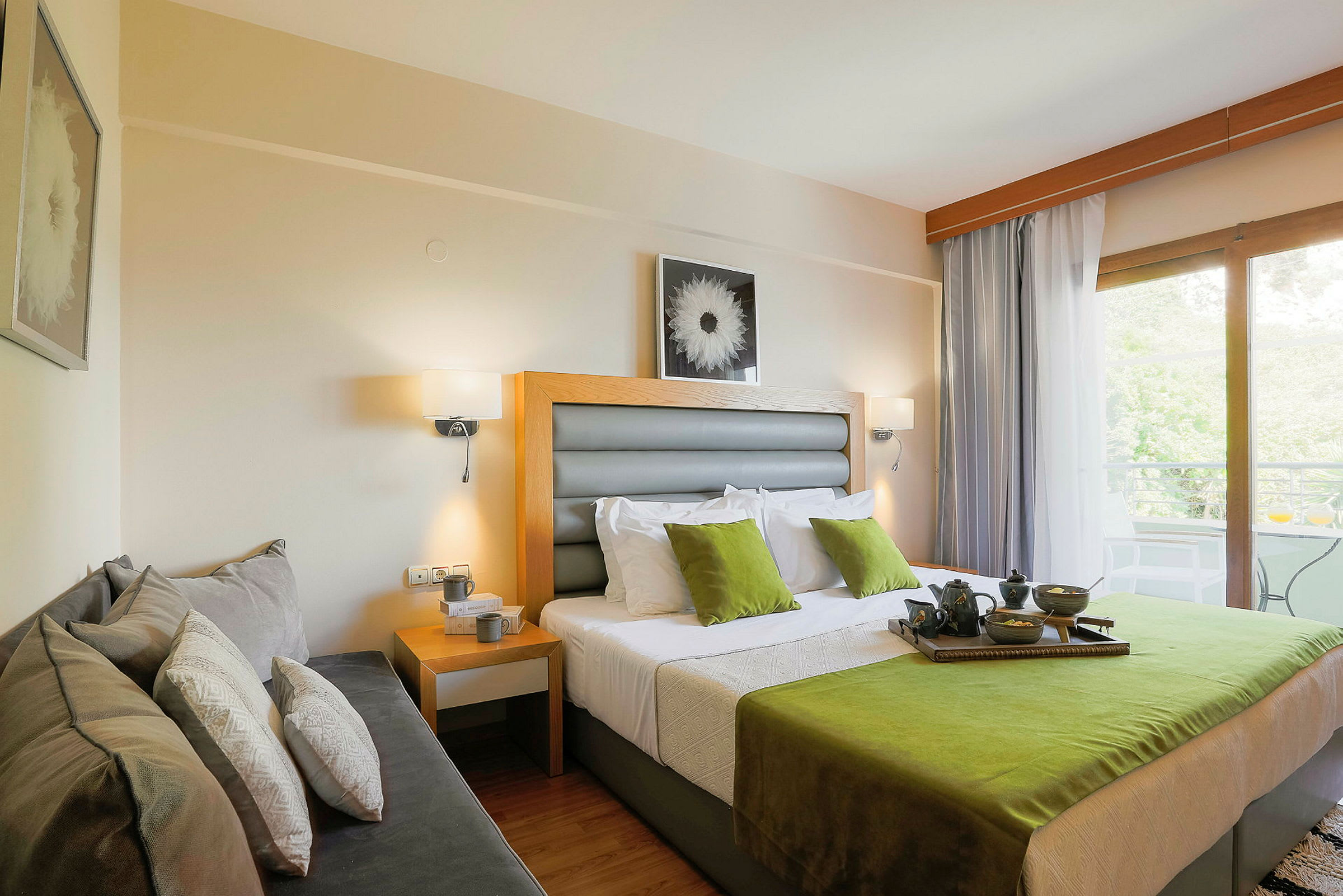 Et hotelværelse med en dobbeltseng med grønne og hvide detaljer, en sofa ved siden af og et vindue, der fører ud til en altan med udsigt. (AI-genereret tekst)