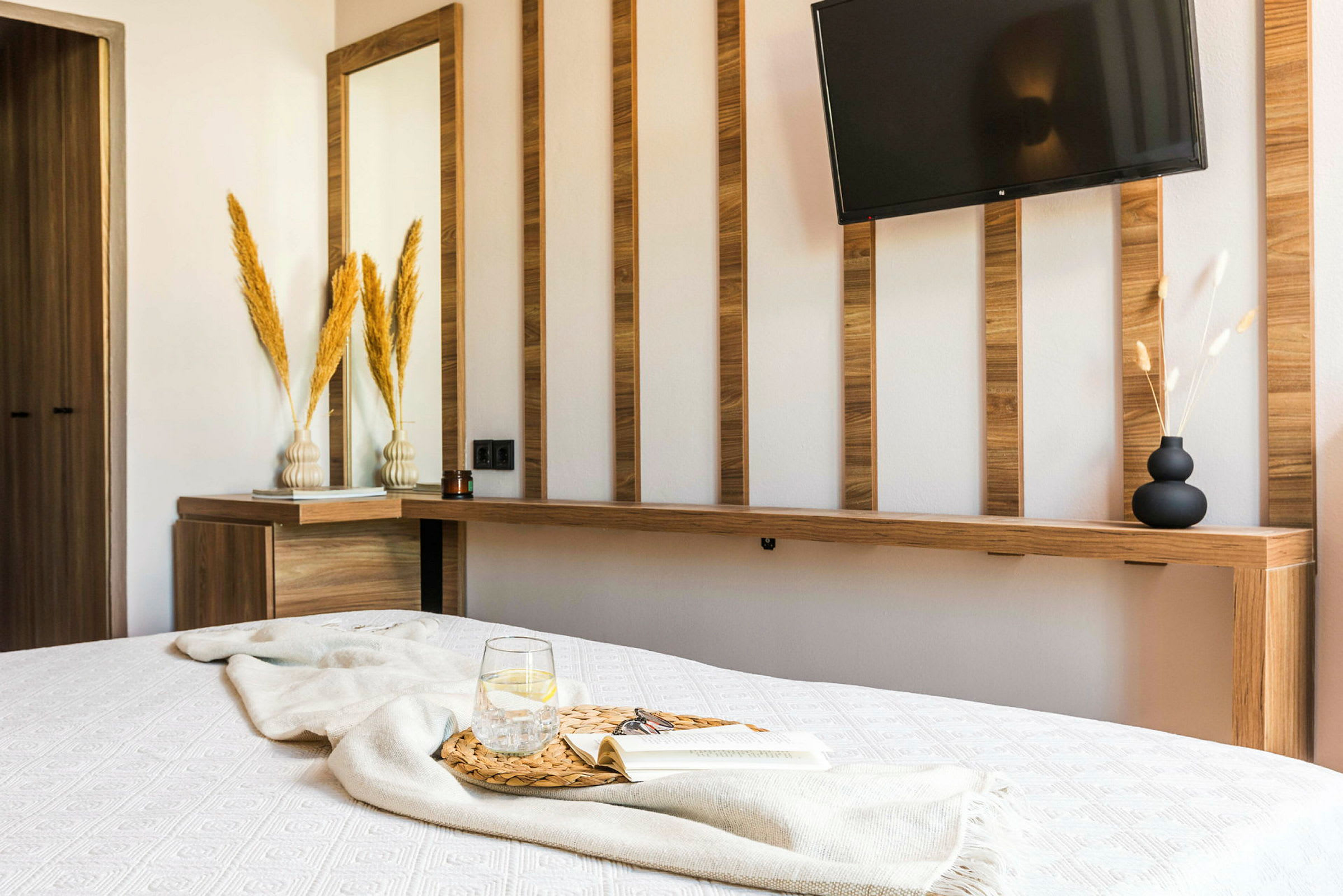 Et moderne designet hotelværelse med et hvidt sengetæppe, et glas vand og en bog på en træbakke samt en væg med træpaneler, et spejl og et fjernsyn. (AI-genereret tekst)