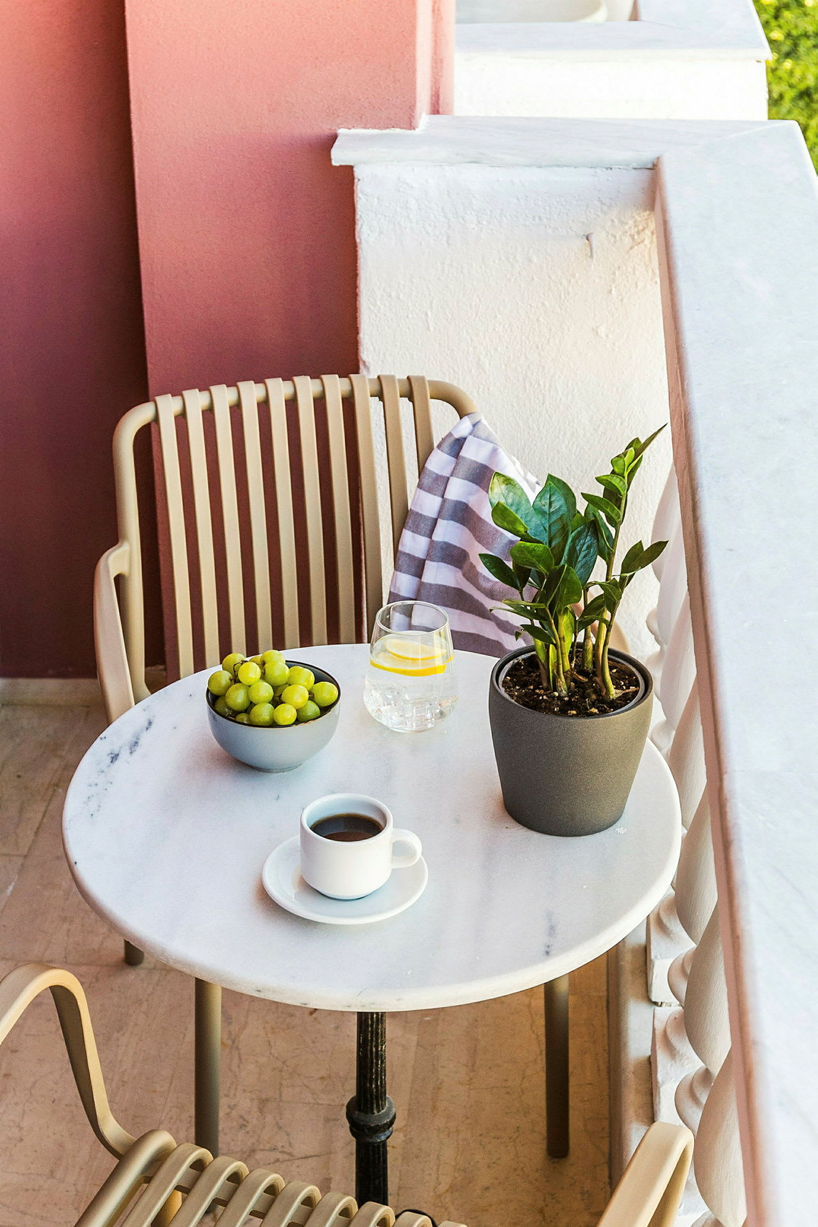 En balkon med et bord med en kaffekop, et glas vand, en skål med vindruer og en grøn plante. (AI-genereret tekst)