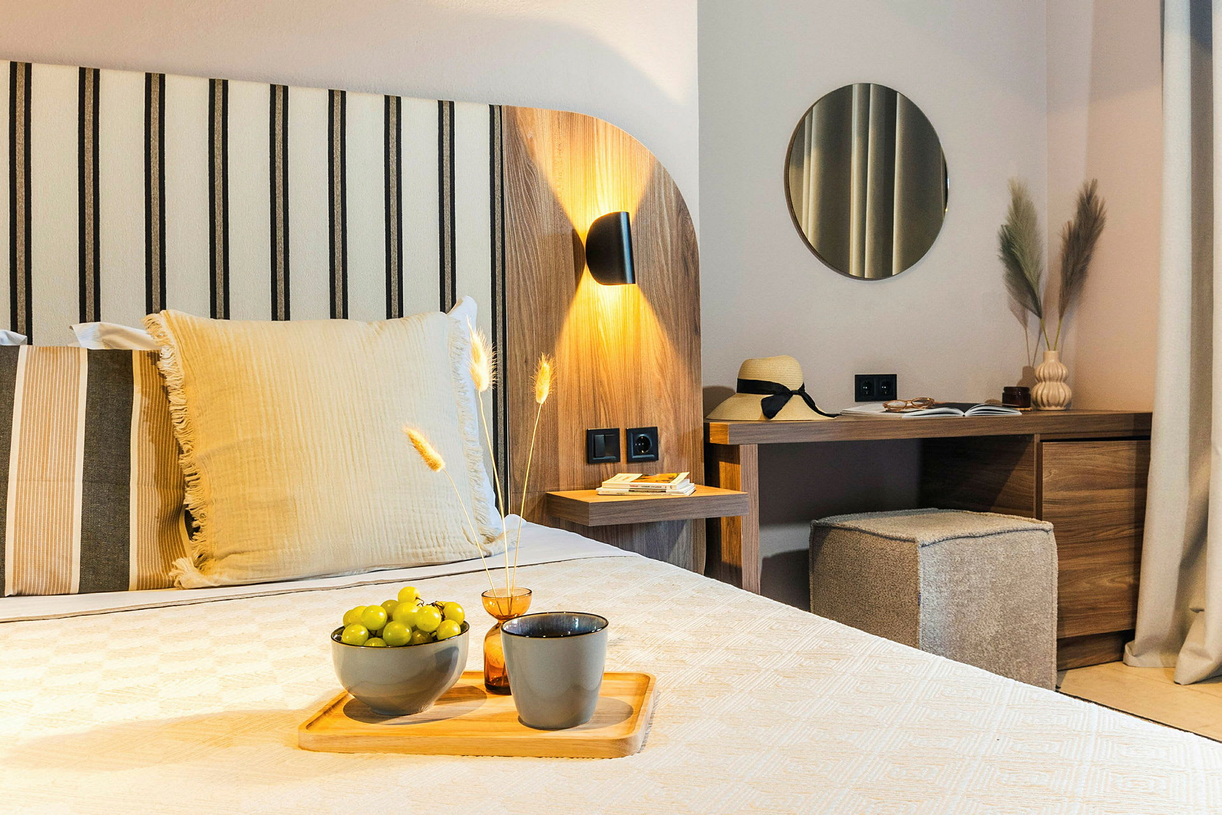 Et moderne hotelværelse med trædekoration, en seng med dekorative puder, et skrivebord med spejl og dekorative genstande, og grønne druer på en tallerken. (AI-genereret tekst)