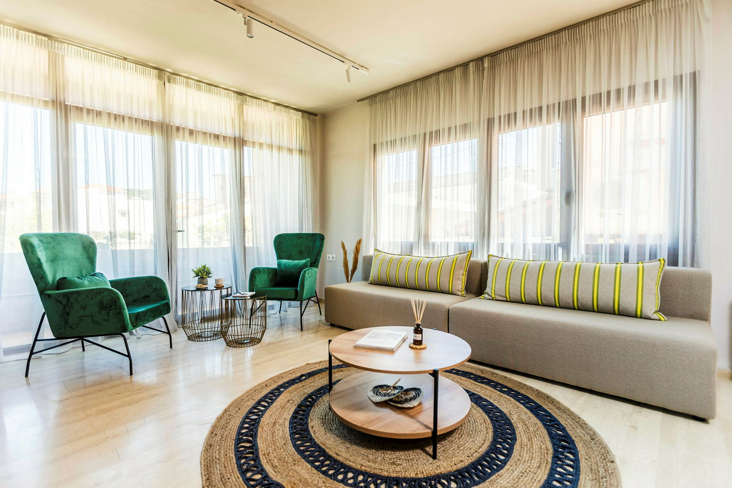Et lyst opholdsrum med store vinduer, to grønne lænestole, en beige sofa med stribede puder og et rundt sofabord med dekorationer. (AI-genereret tekst)