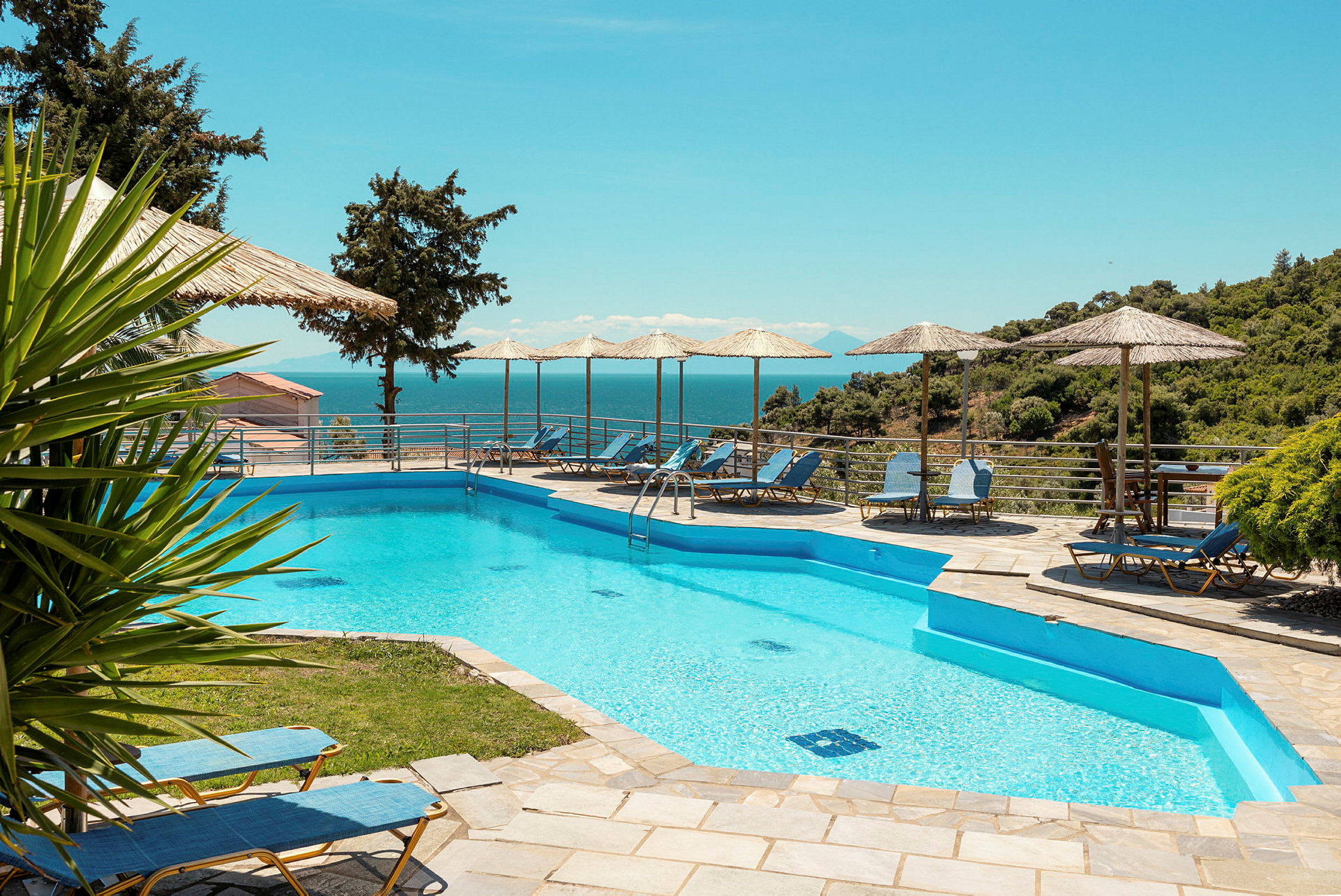 Udendørs swimmingpool med liggestole og parasoller ved siden. I baggrunden ses havet og frodigt landskab. (AI-genereret tekst)
