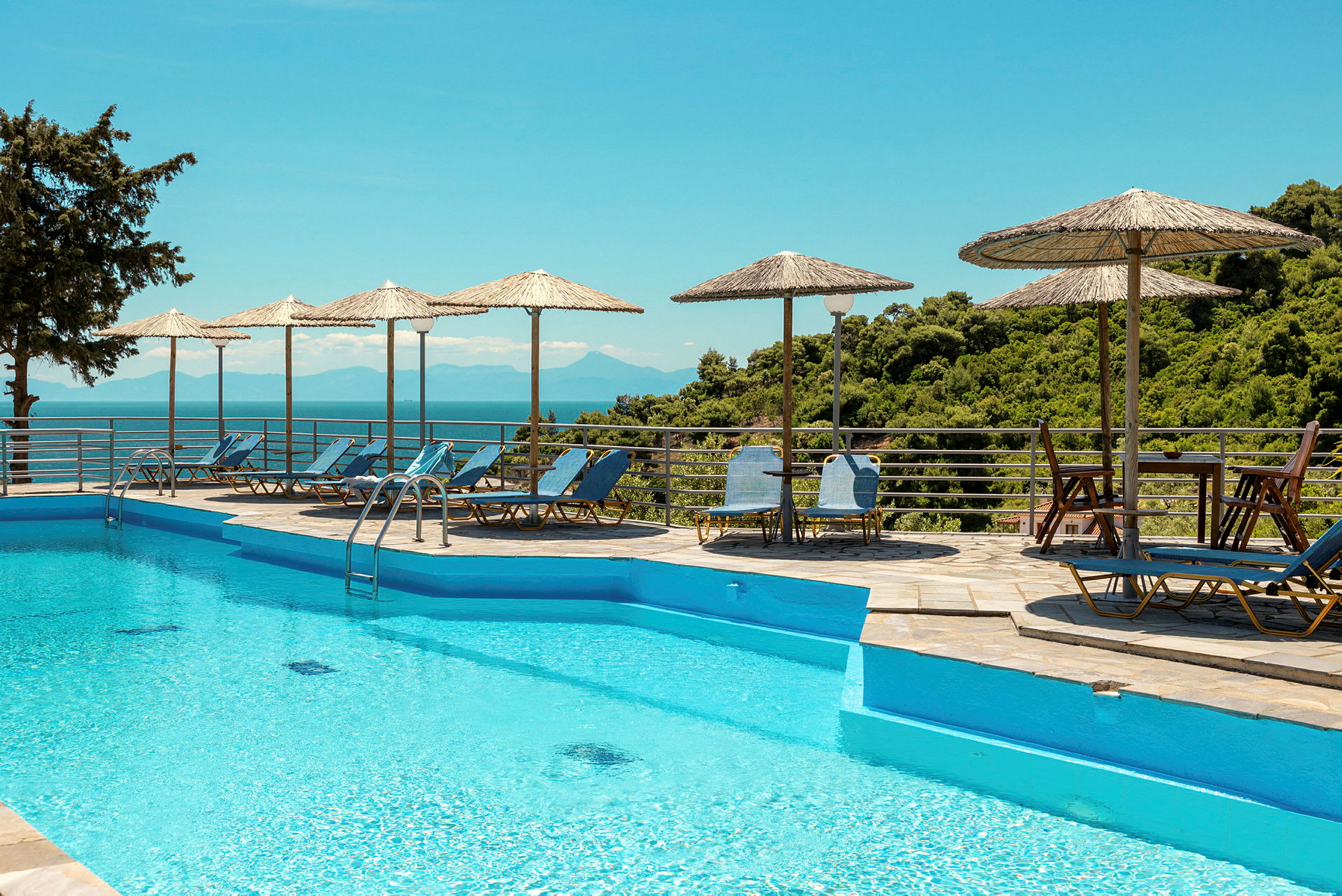 Pool med blåt vand, omgivet af liggestole og parasoller med udsigt over natur og hav. (AI-genereret tekst)