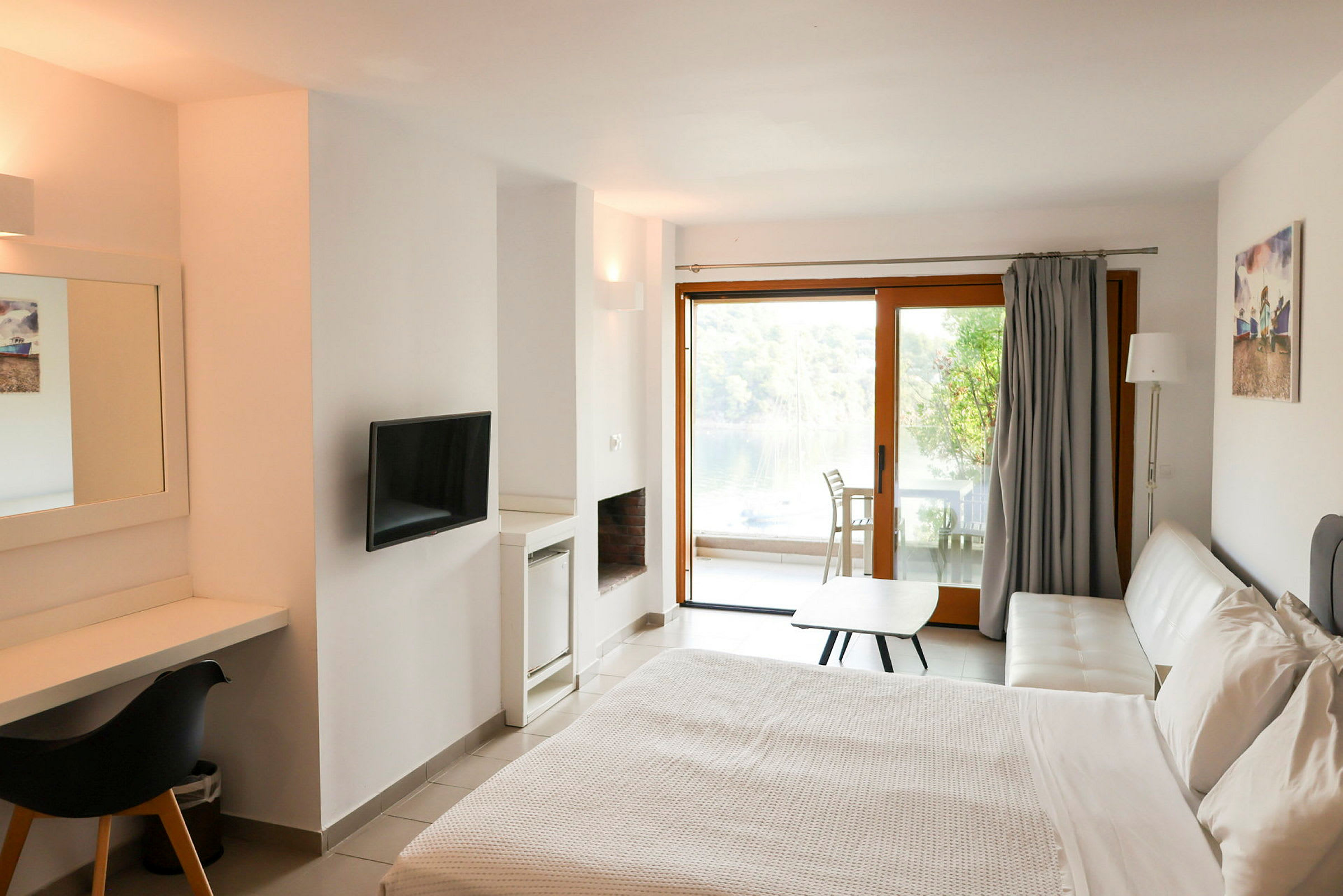 Et moderne hotelværelse med en dobbeltseng, skrivebord, TV og en hvid sofa. I baggrunden ses en glasdør, der fører til en møbleret terrasse med udsigt over natur og vand. (AI-genereret tekst)