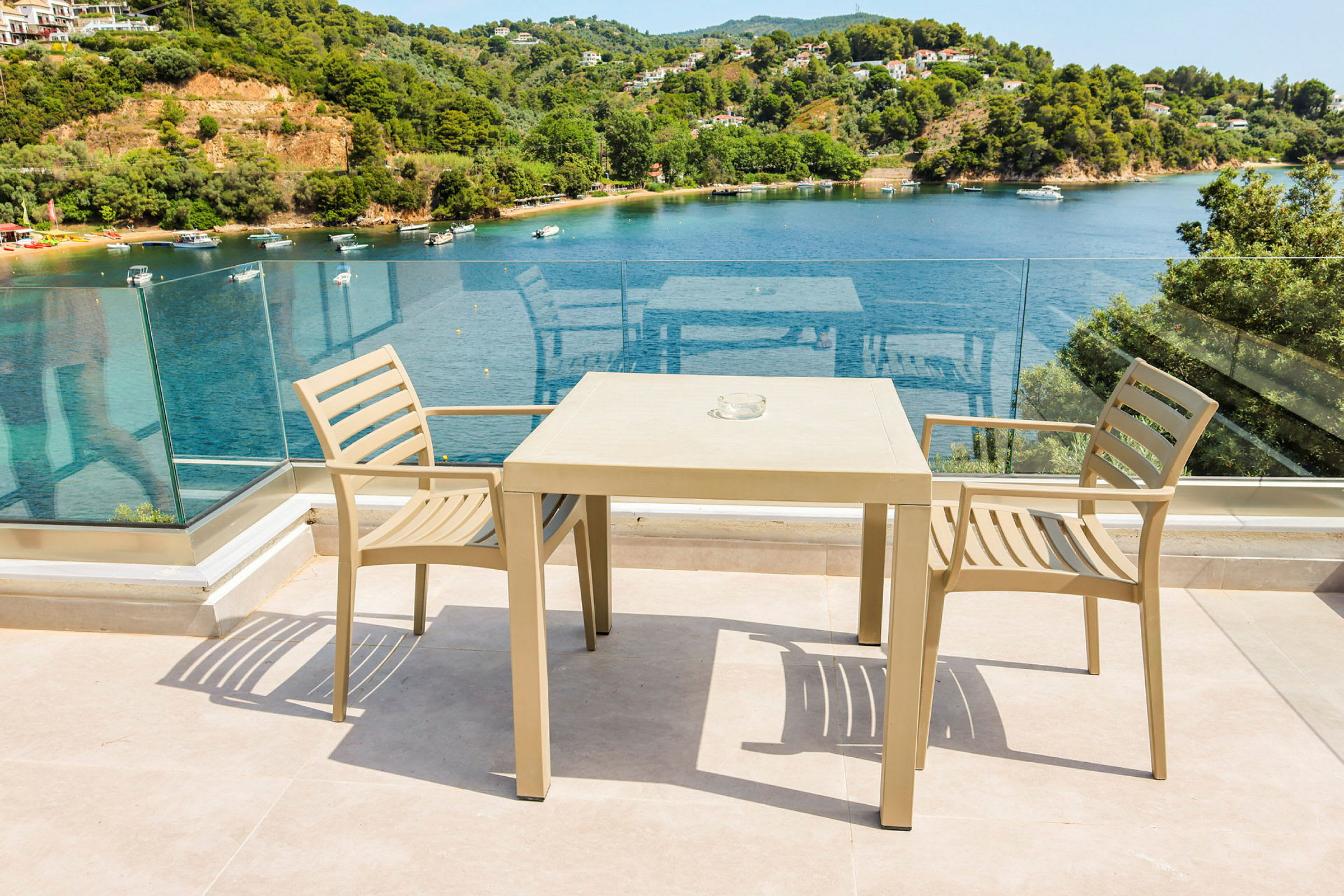 To beige stole og et bord på en terrasse med glasrækværk og udsigt over en bugt med både og grøntområde. (AI-genereret tekst)