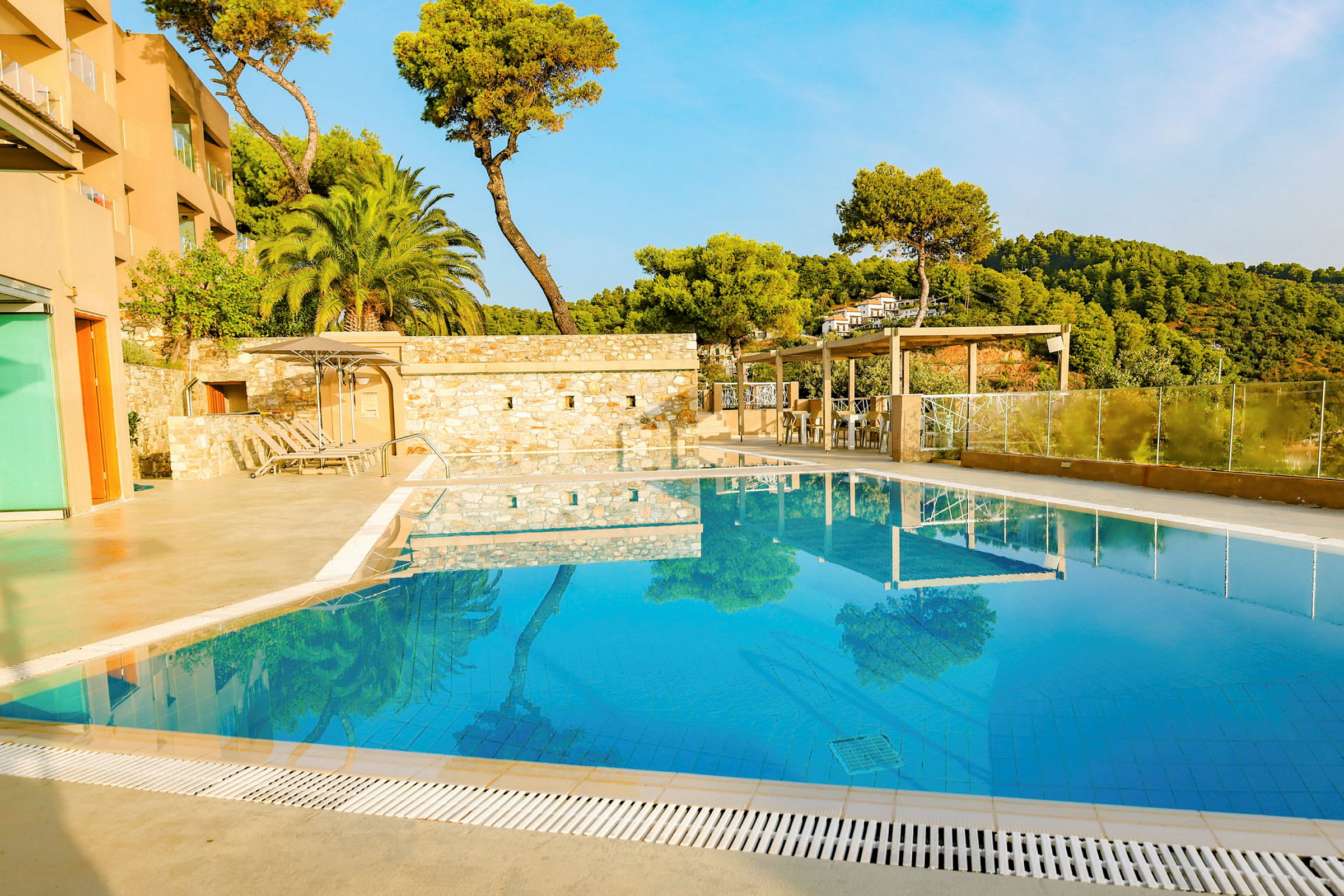 Udendørs swimmingpool ved hotelbygning omgivet af grønne områder og træer. (AI-genereret tekst)