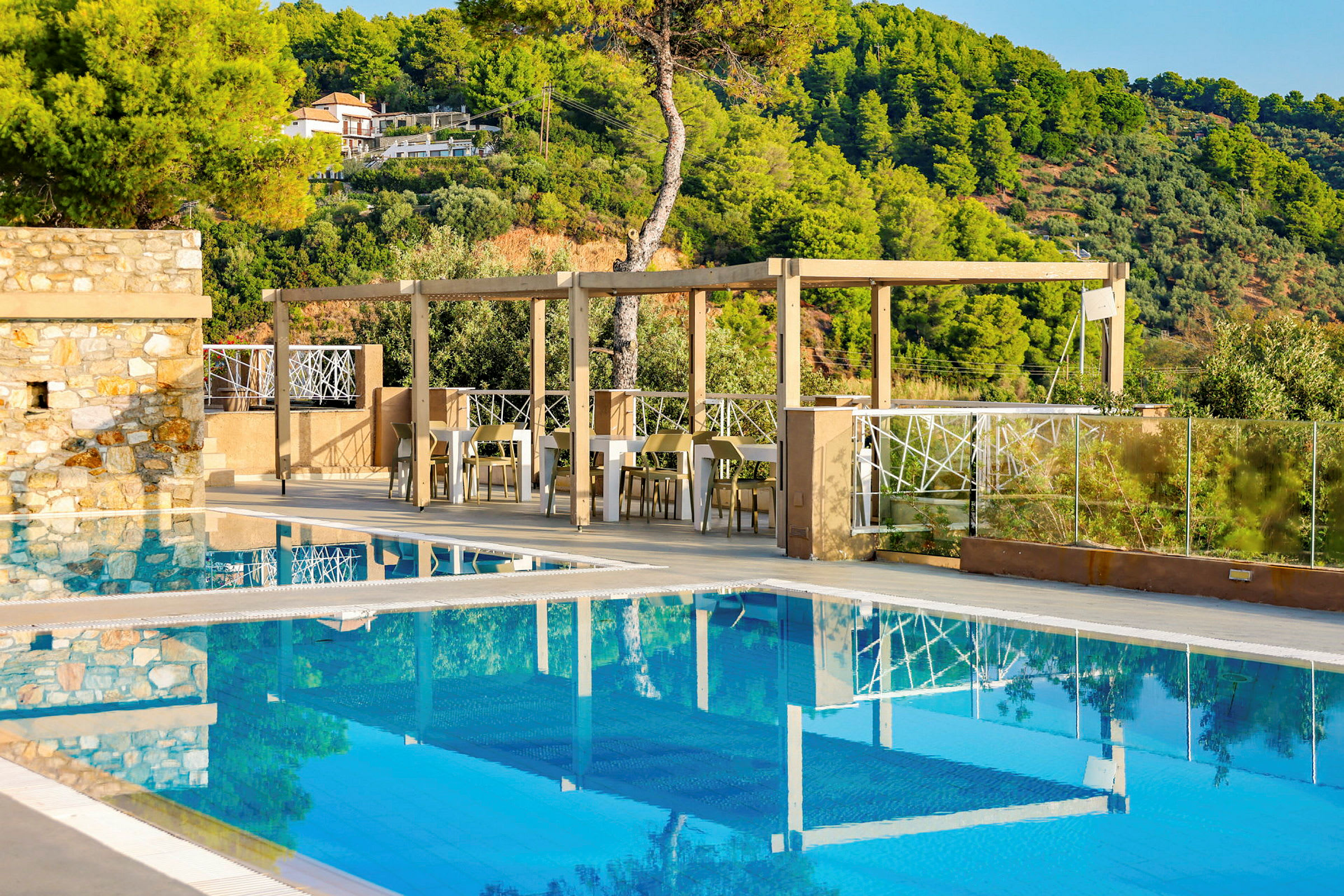 Udendørs pool med omgivelser af grønne områder og siddepladser under pergola. (AI-genereret tekst)