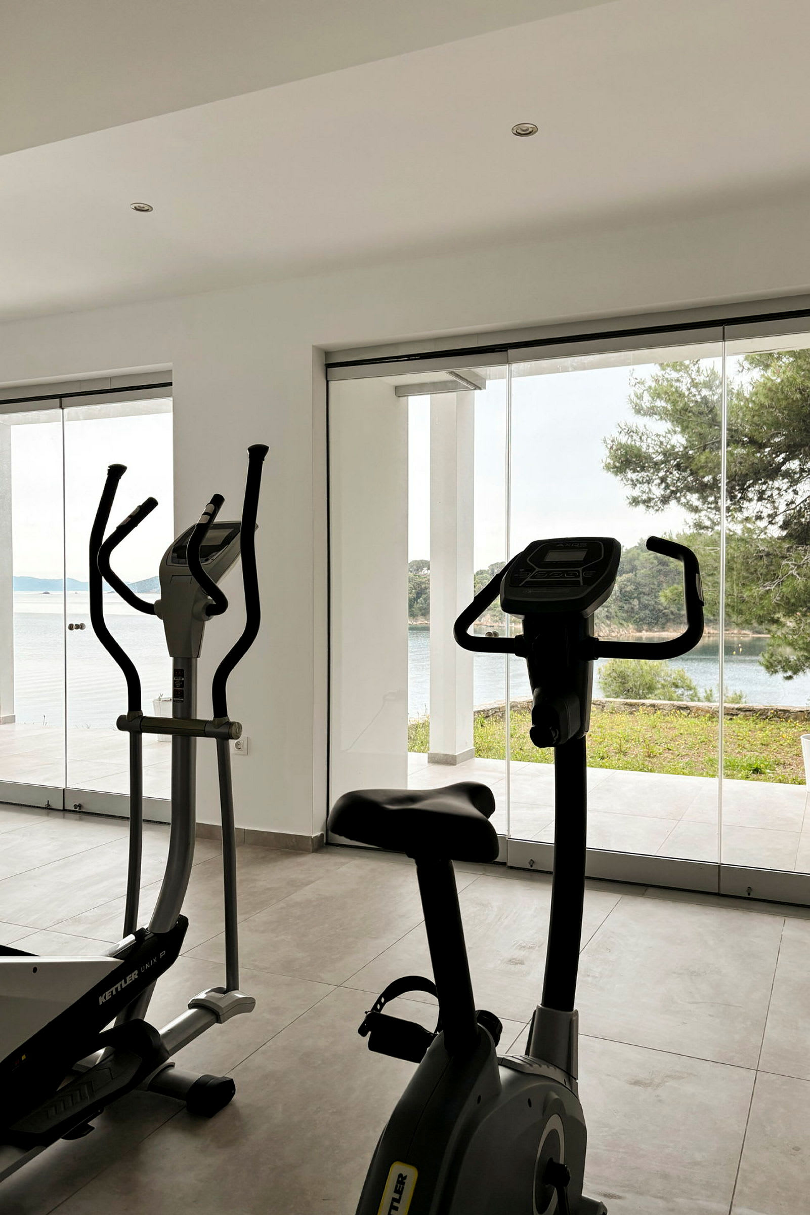 Et fitnesscenter med træningsmaskiner, herunder motionscykel og crosstrainer, med store vinduer, der giver udsigt til natur og hav. (AI-genereret tekst)