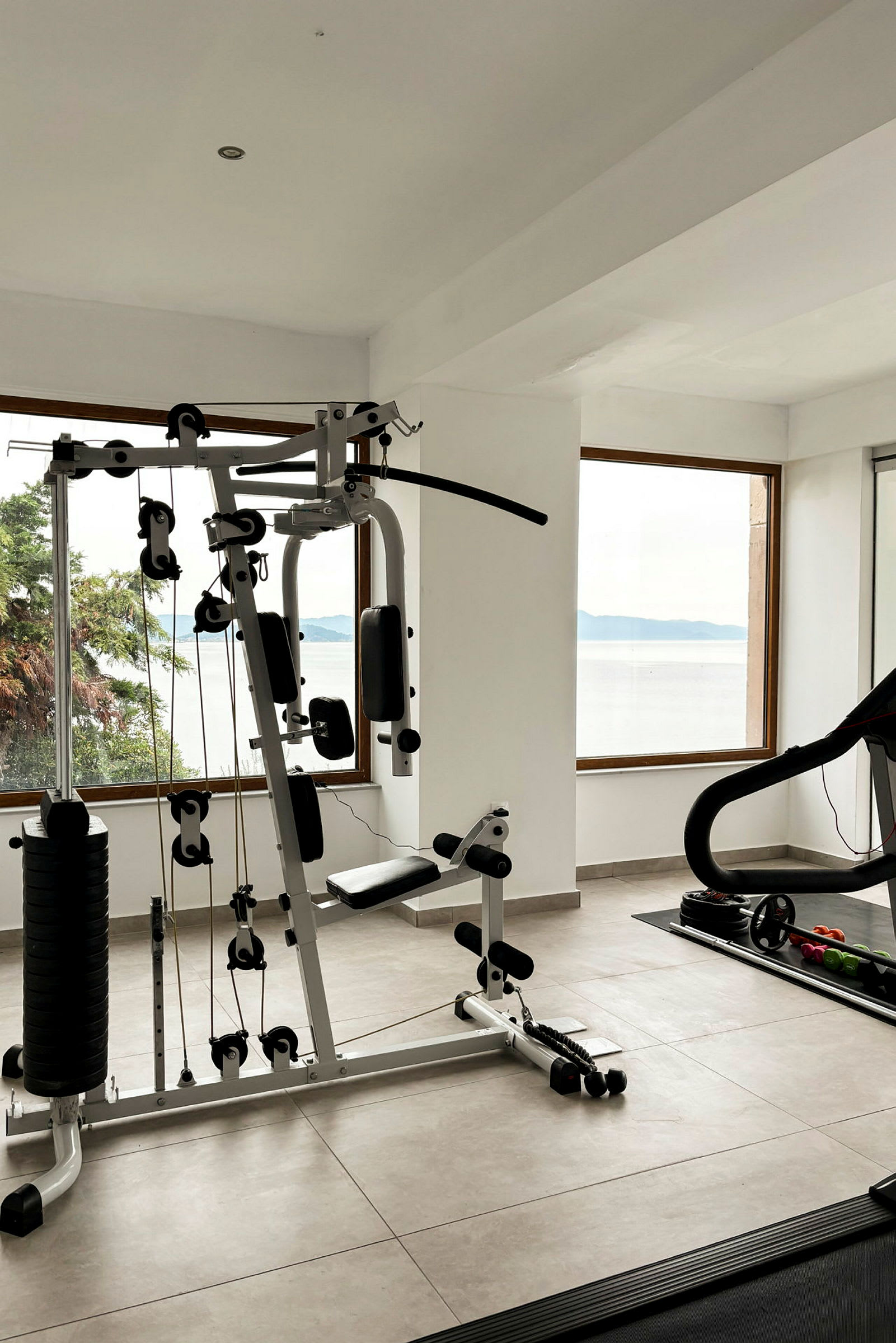 Et træningsrum med udstyr, herunder et multigym og et løbebånd, med vinduer med udsigt over havet. (AI-genereret tekst)