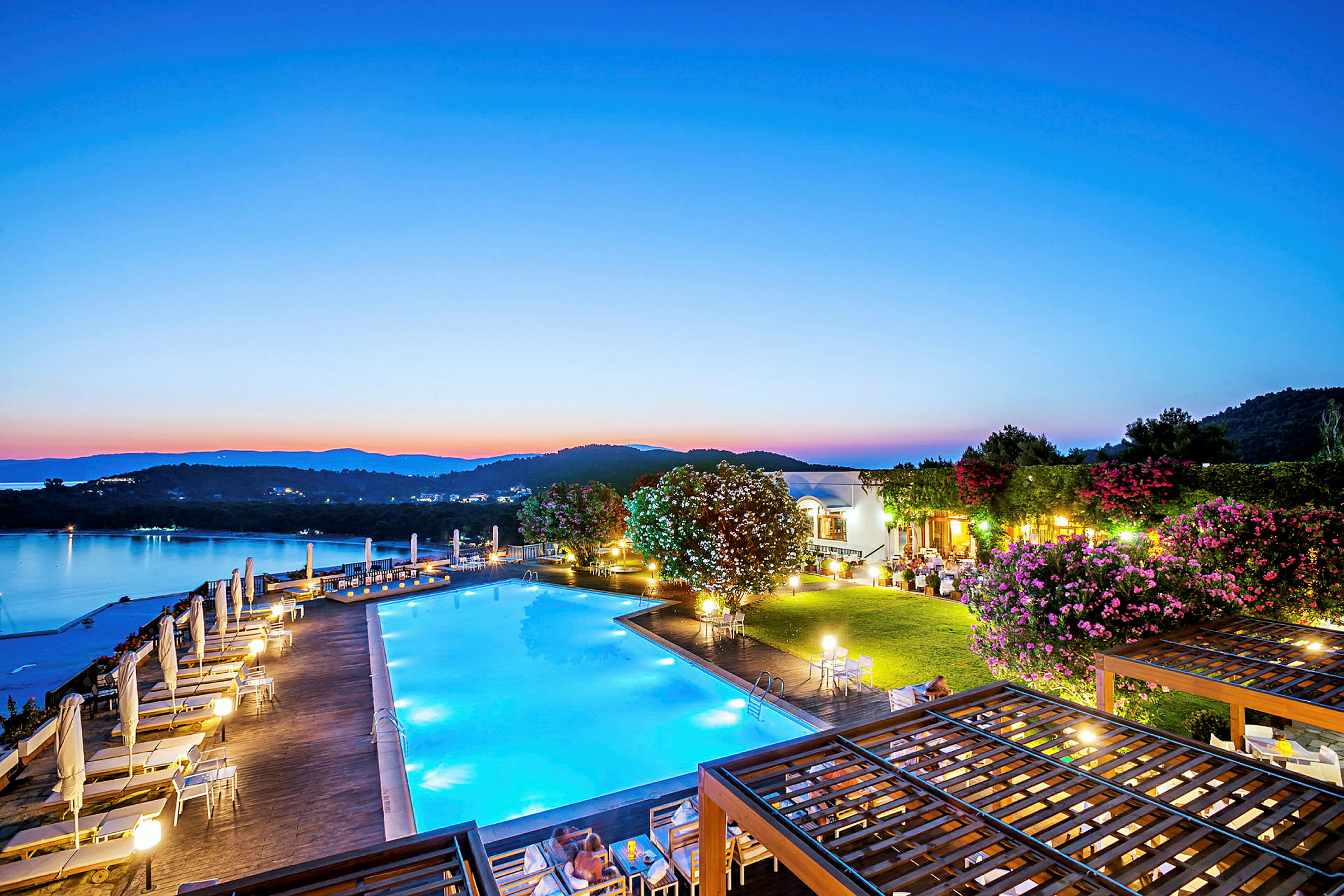 En pool ved en resort med udsigt over en rolig sø og omkringliggende bakker i skumringen. (AI-genereret tekst)