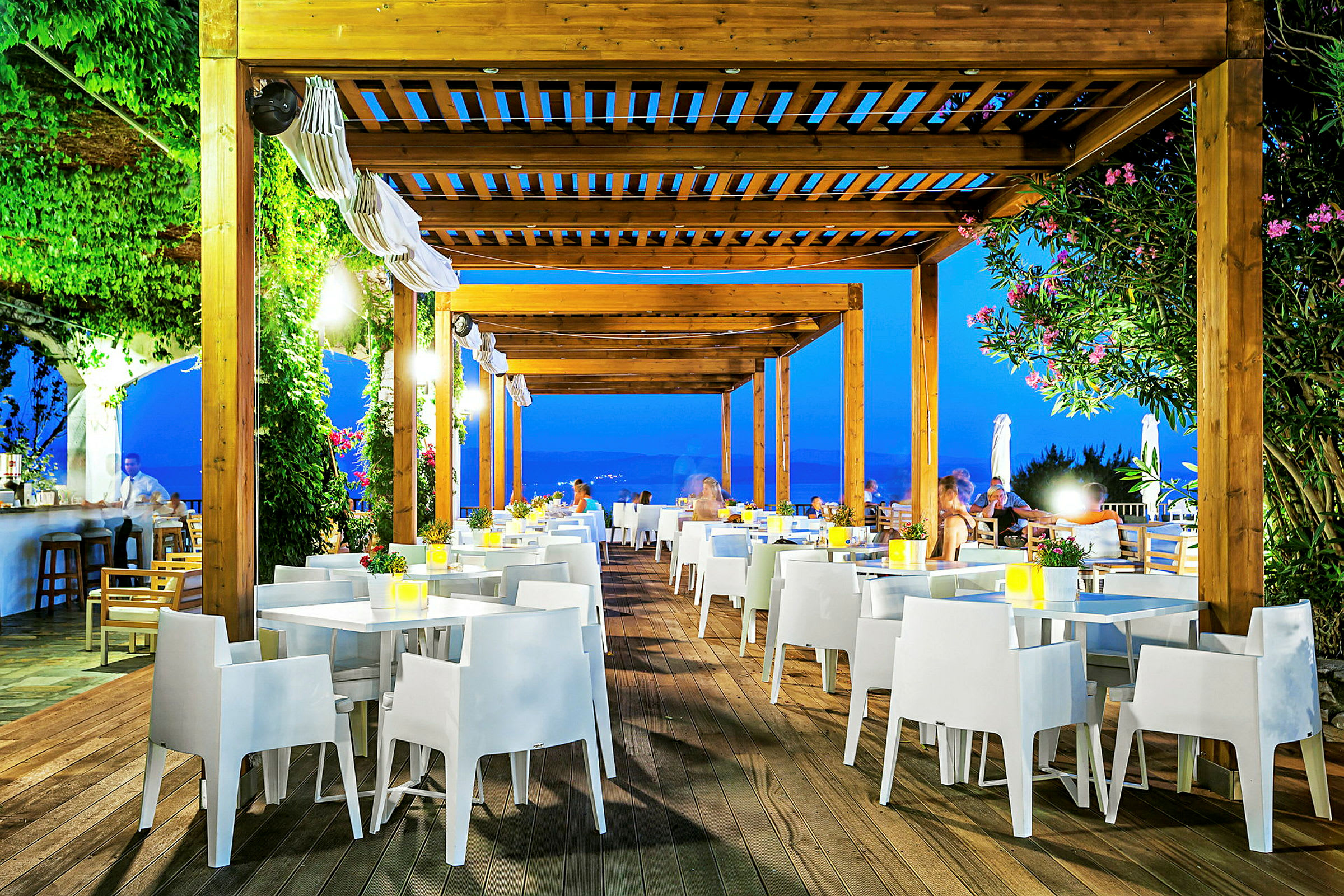 Udendørsrestaurant med træpergola, hvide stole og borde, omgivet af grønt og aftenbelysning. (AI-genereret tekst)