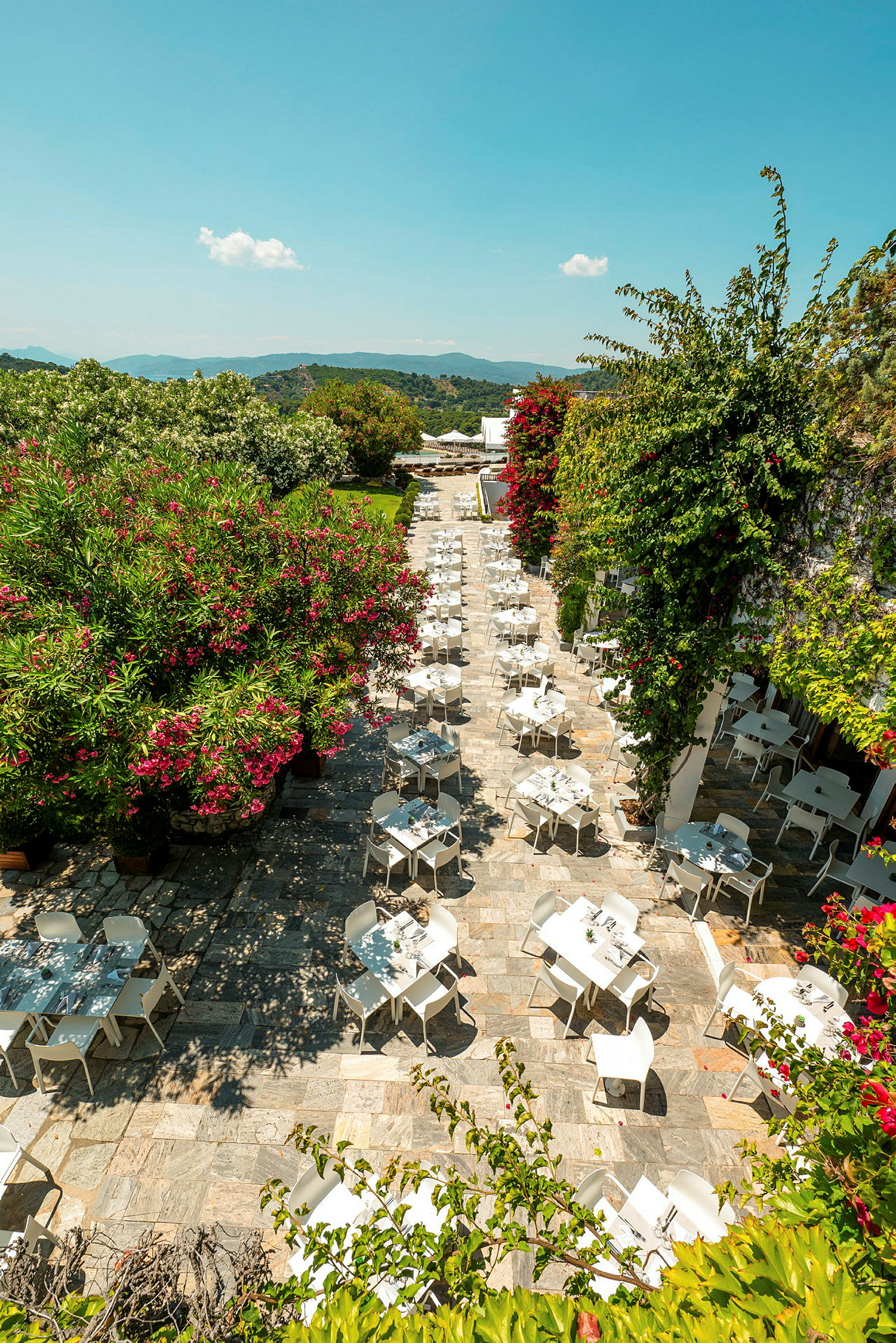 Udendørs terrasse med hvide borde og stole omgivet af frodig vegetation og blomster. (AI-genereret tekst)