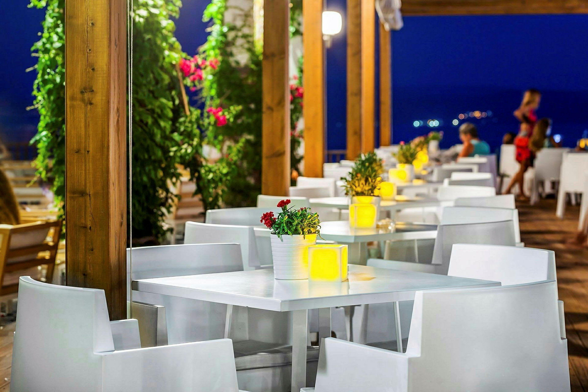 En udendørs restaurant med hvide borde og stole, dekoreret med blomster og lys om aftenen. (AI-genereret tekst)
