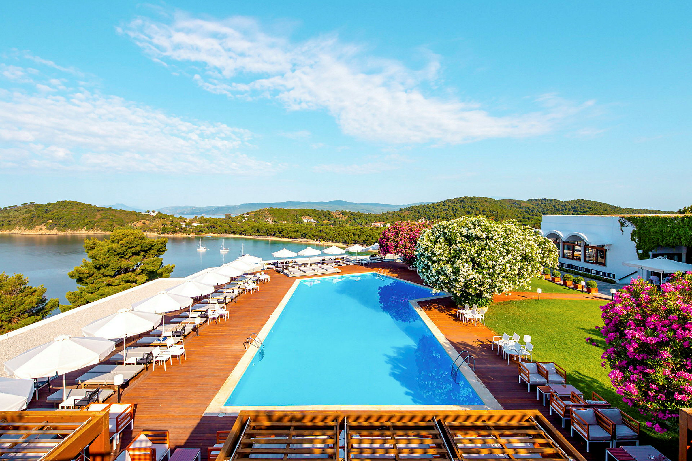 Udendørs område med pool og terrasse ved havet, omgivet af frodig vegetation og bygninger i baggrunden. (AI-genereret tekst)
