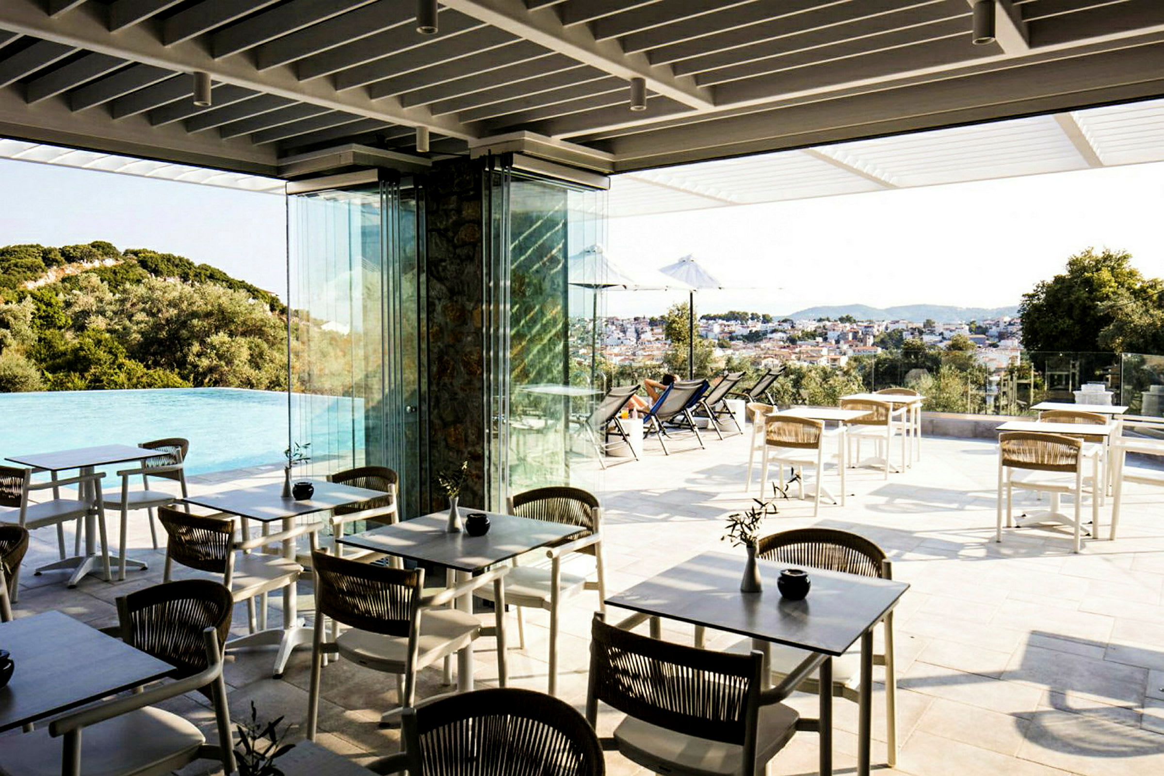 En udendørs terrasse med borde og stole nær en infinitypool med udsigt over et landskab og en by. (AI-genereret tekst)