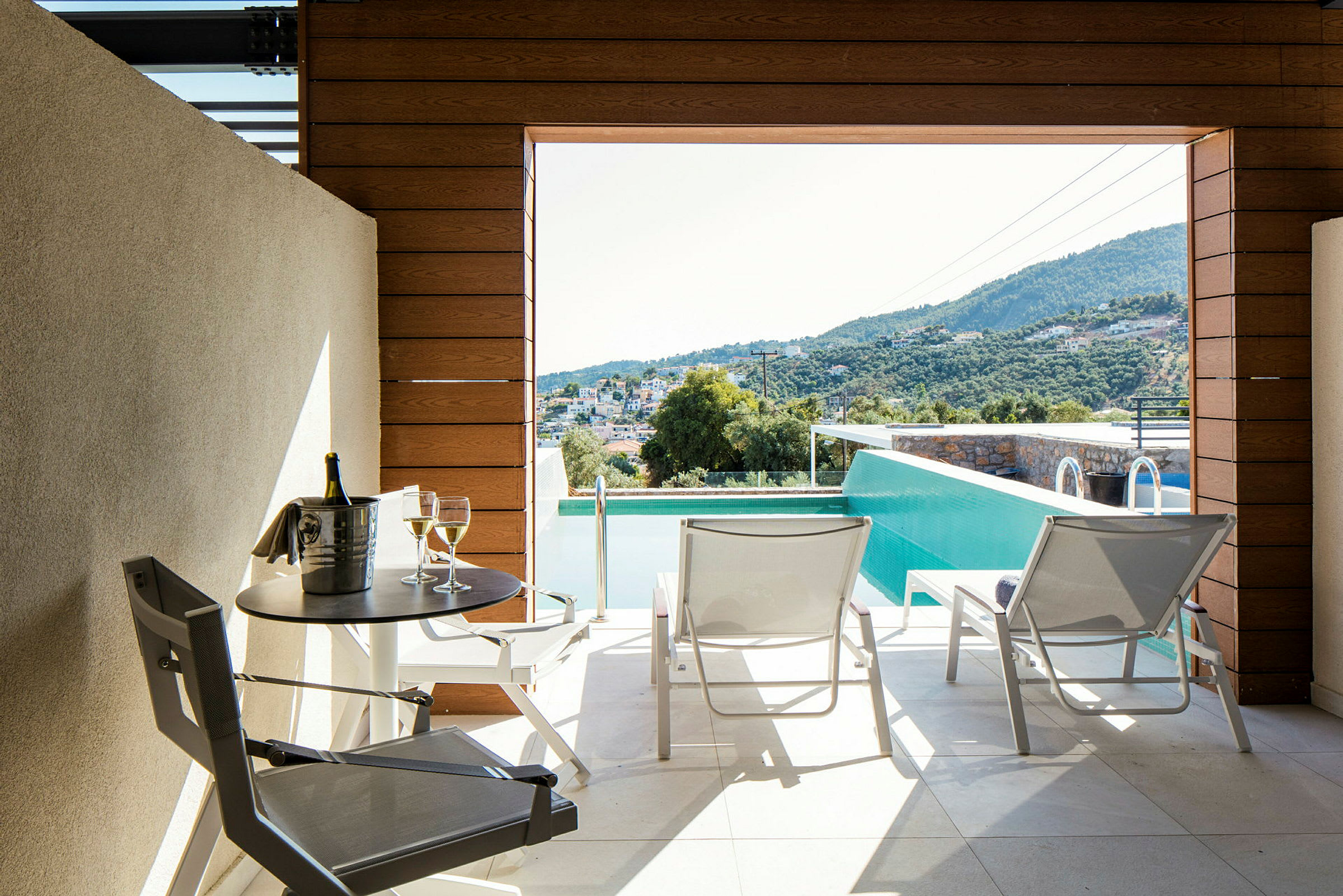 Terrasse med udsigt over poolen og landskabet, med to liggestole og et bord med en flaske og to glas. (AI-genereret tekst)
