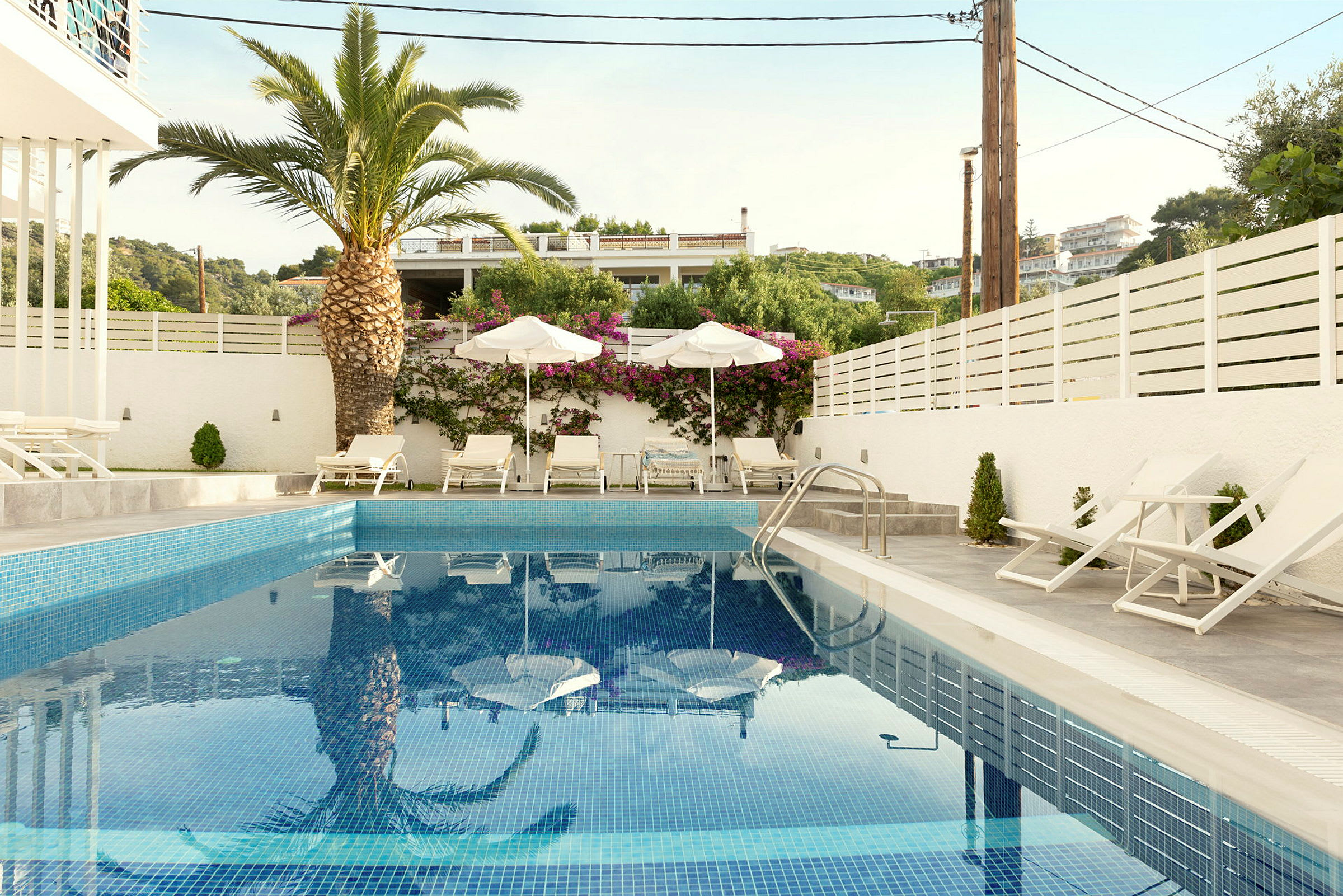 Udendørs pool med liggestole og parasoller, omgivet af en hvid mur og grønne planter. (AI-genereret tekst)