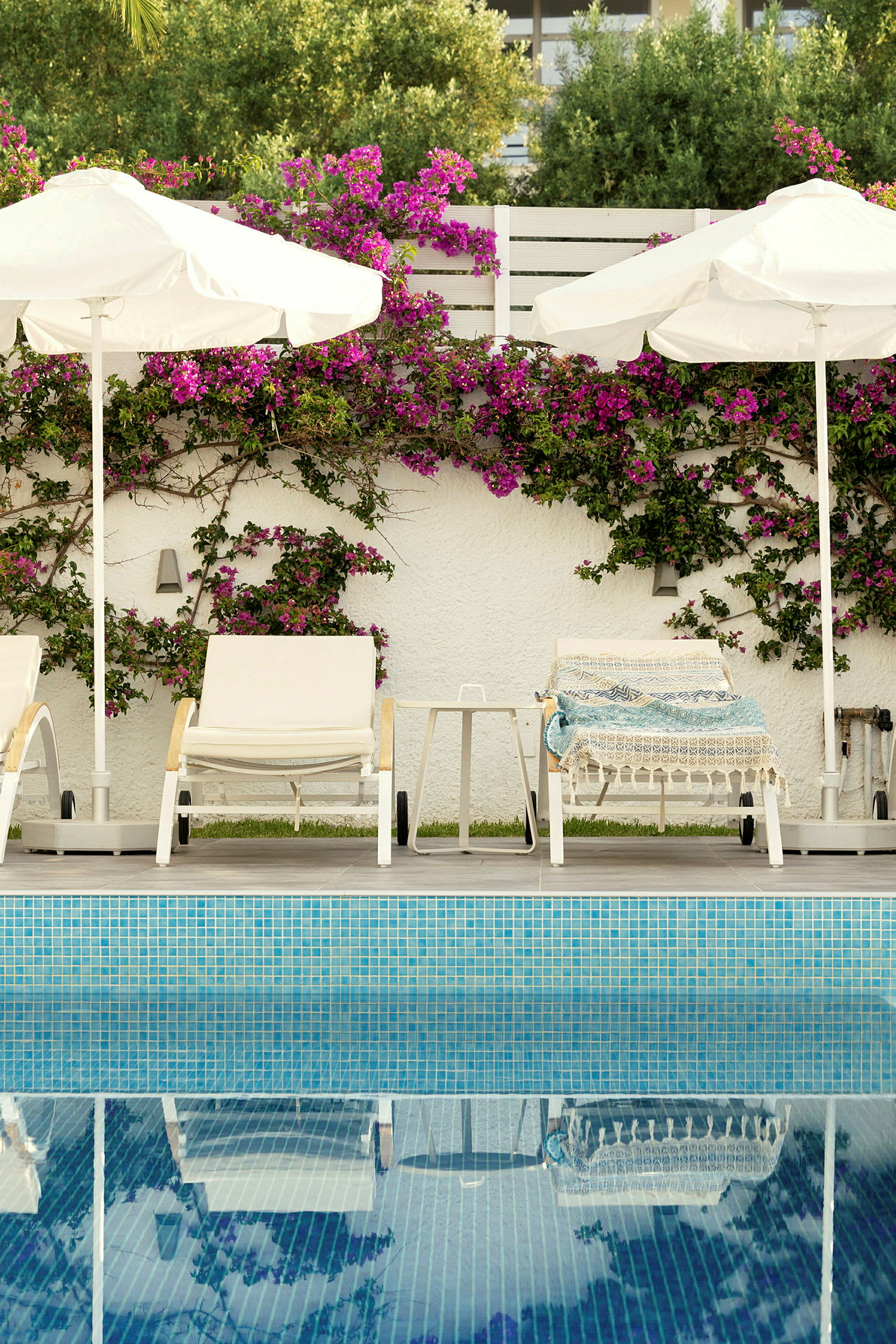 Billede af liggestole og parasoller ved en pool, med en væg dekoreret med blomster i baggrunden. (AI-genereret tekst)