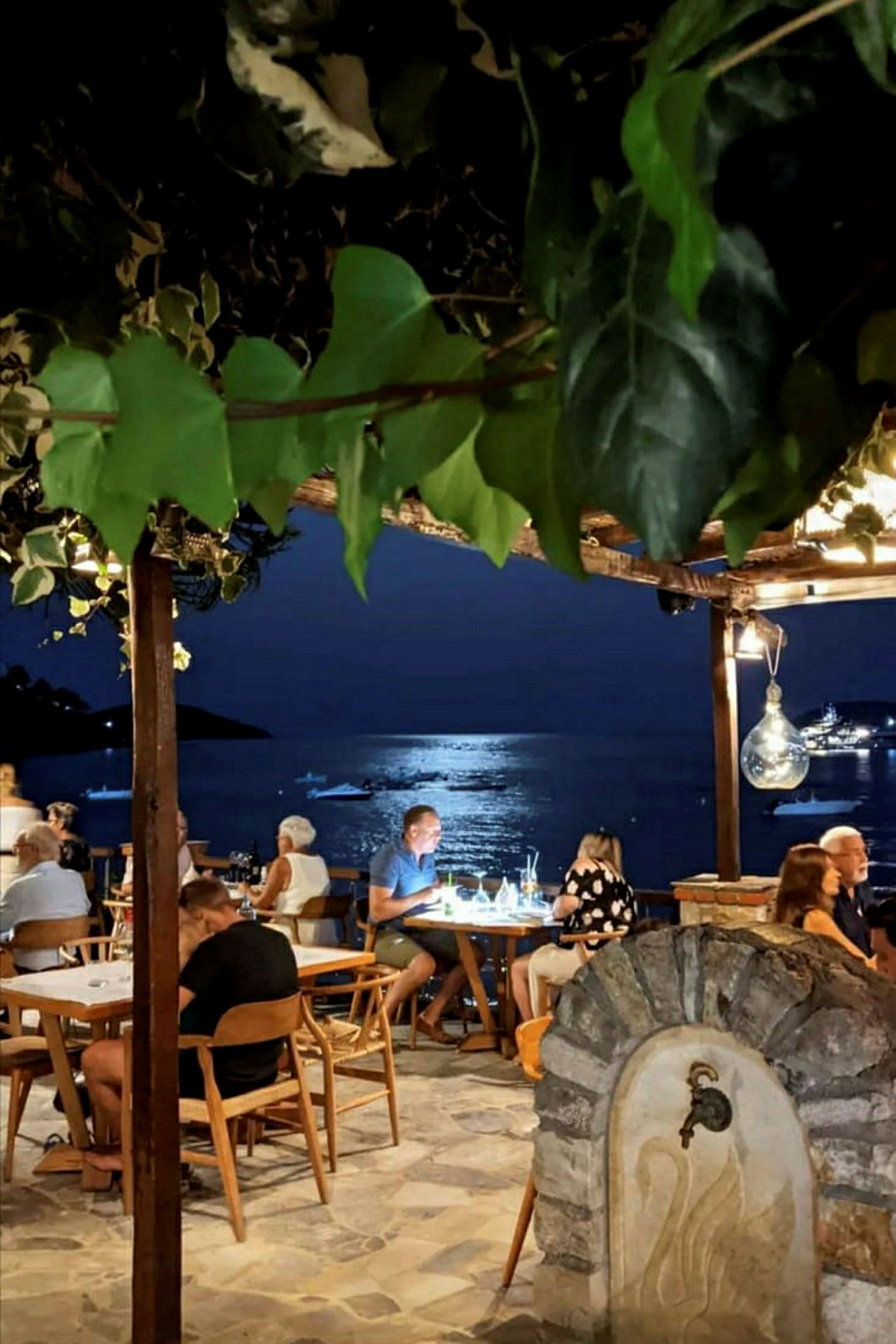 Udendørs servering ved havet med lys og både synlige i baggrunden. (AI-genereret tekst)