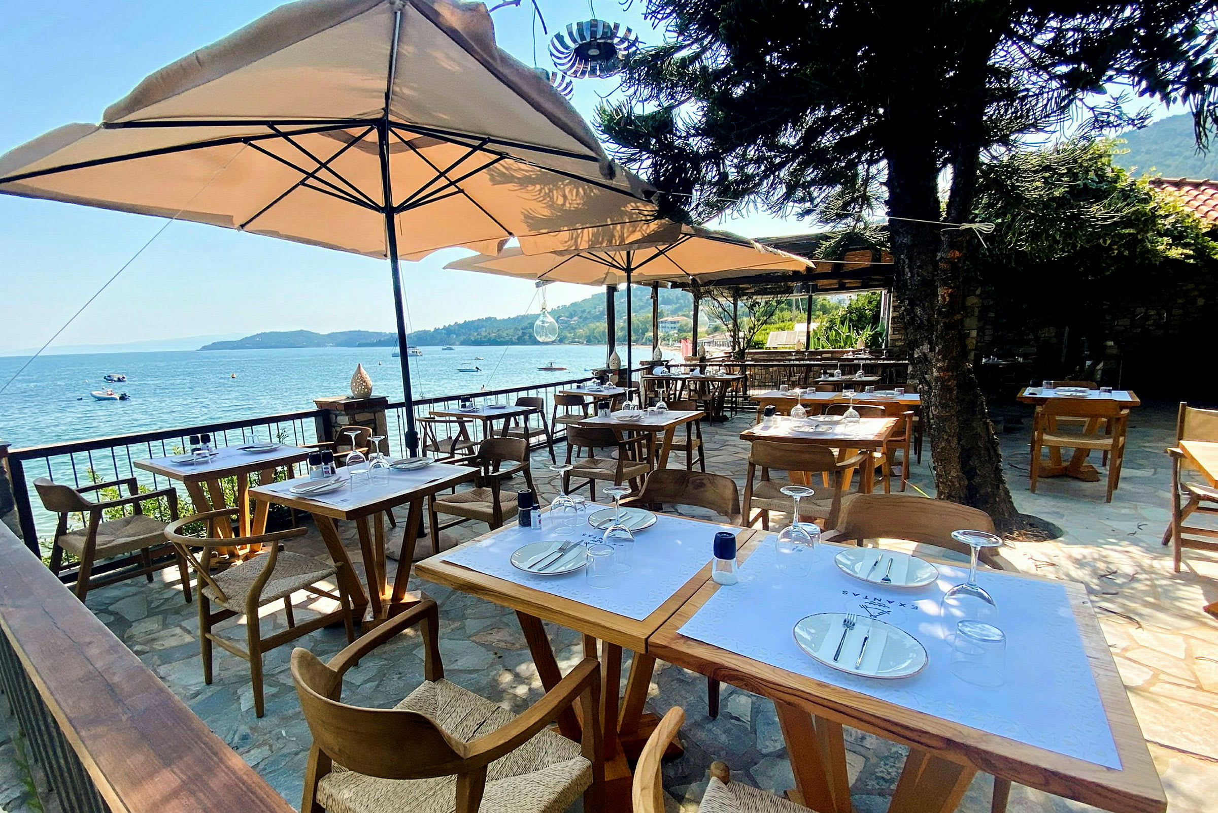 Billede af en udendørs restaurant med borde og stole under parasoller, med udsigt til havet i baggrunden. (AI-genereret tekst)