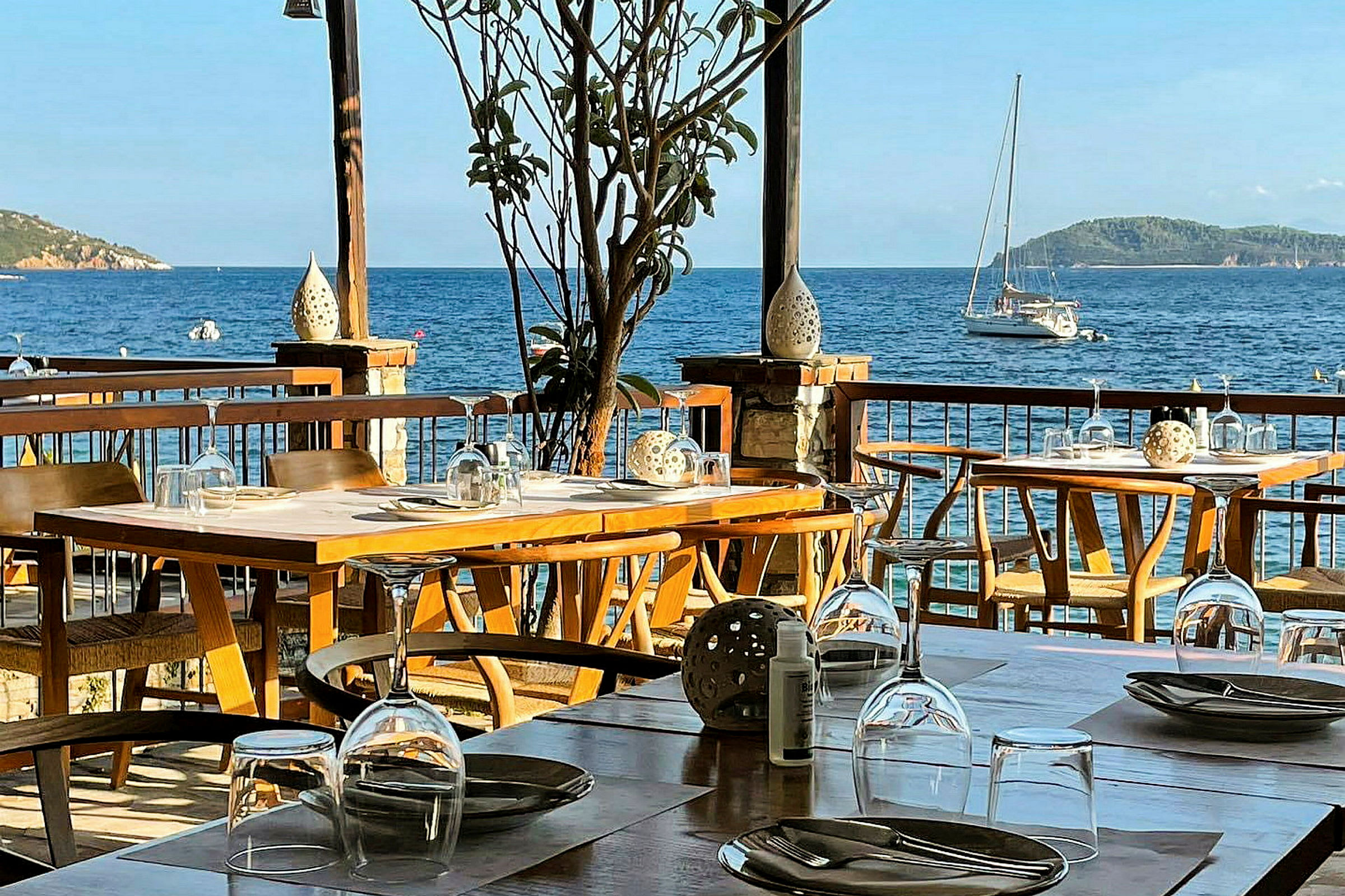 Udendørs serveringsområde med dækkede borde og udsigt over havet med en sejlbåd. (AI-genereret tekst)