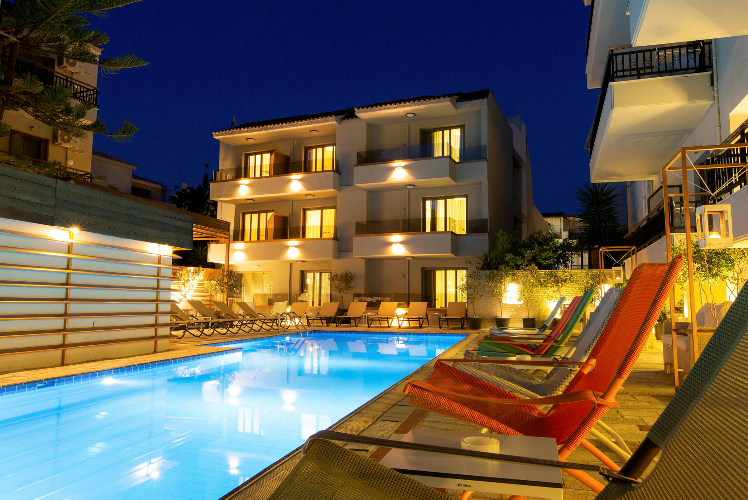 Billedet viser en oplyst hotelbygning og en udendørs pool omgivet af solsenge og loungestole om aftenen. (AI-genereret tekst)