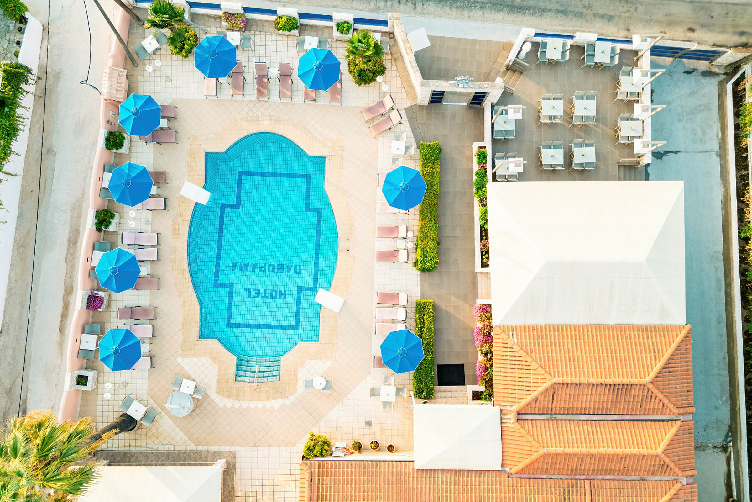 Flyfoto af hotellets poolområde med liggestole og parasoller. (AI-genereret tekst)