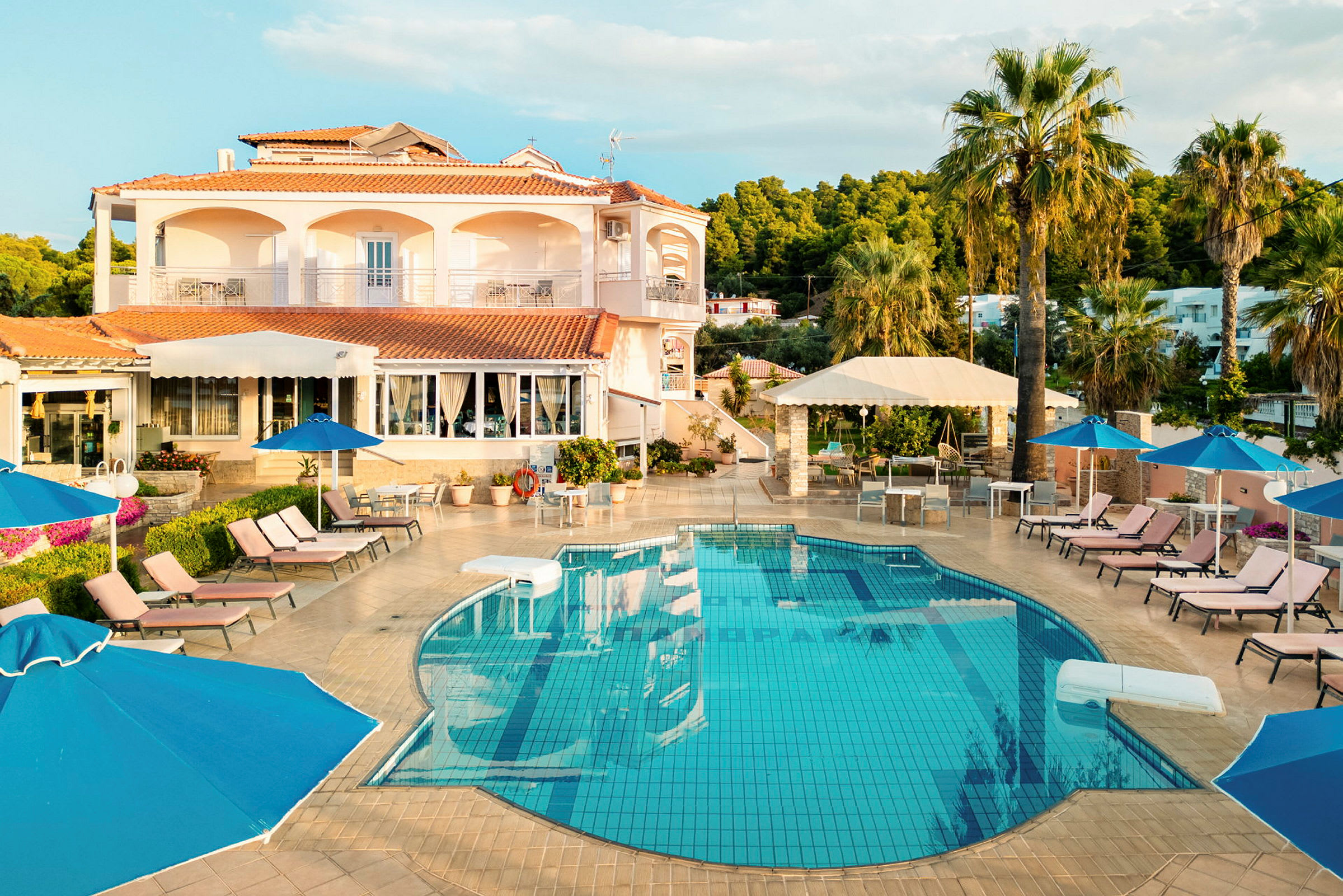 Billede af en udendørs swimmingpool ved et hotel med liggestole, parasoller og palmer omkring samt en bygning i baggrunden. (AI-genereret tekst)
