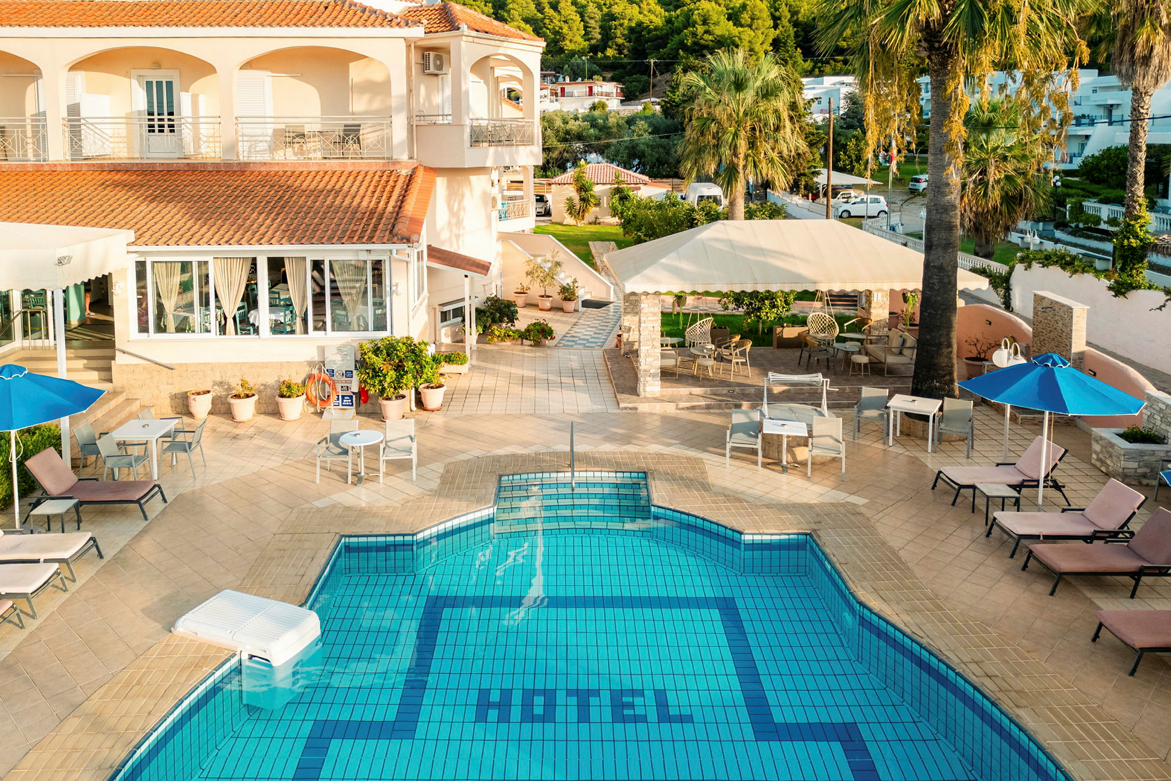 Et hotel med en udendørs pool, liggestole og parasoller med en have i baggrunden. (AI-genereret tekst)