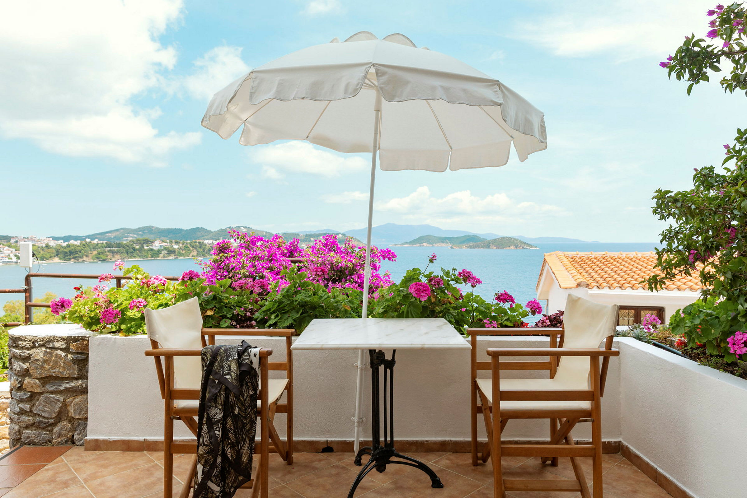 En terrasse med bord, stole, en parasol og blomster, med havudsigt. (AI-genereret tekst)