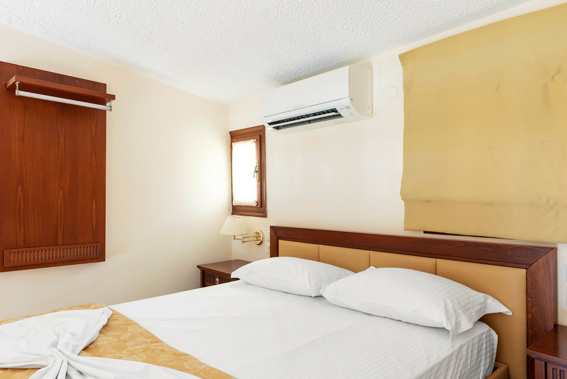 Et hotelværelse med en dobbeltseng, vægmonteret aircondition og et lille vindue. (AI-genereret tekst)