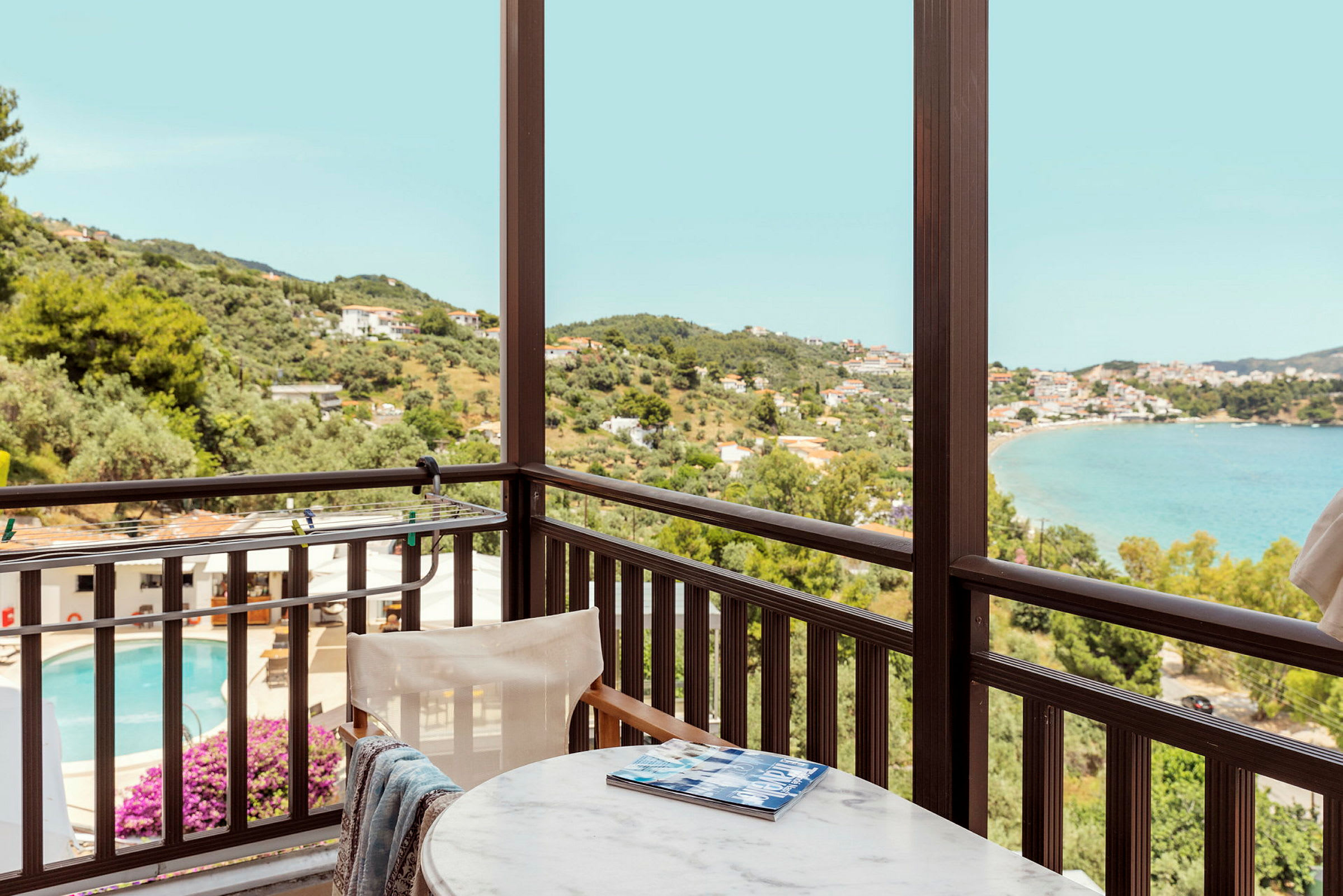 Udsigt fra en balkon med bord og stol mod en pool, vegetation og fjord. (AI-genereret tekst)