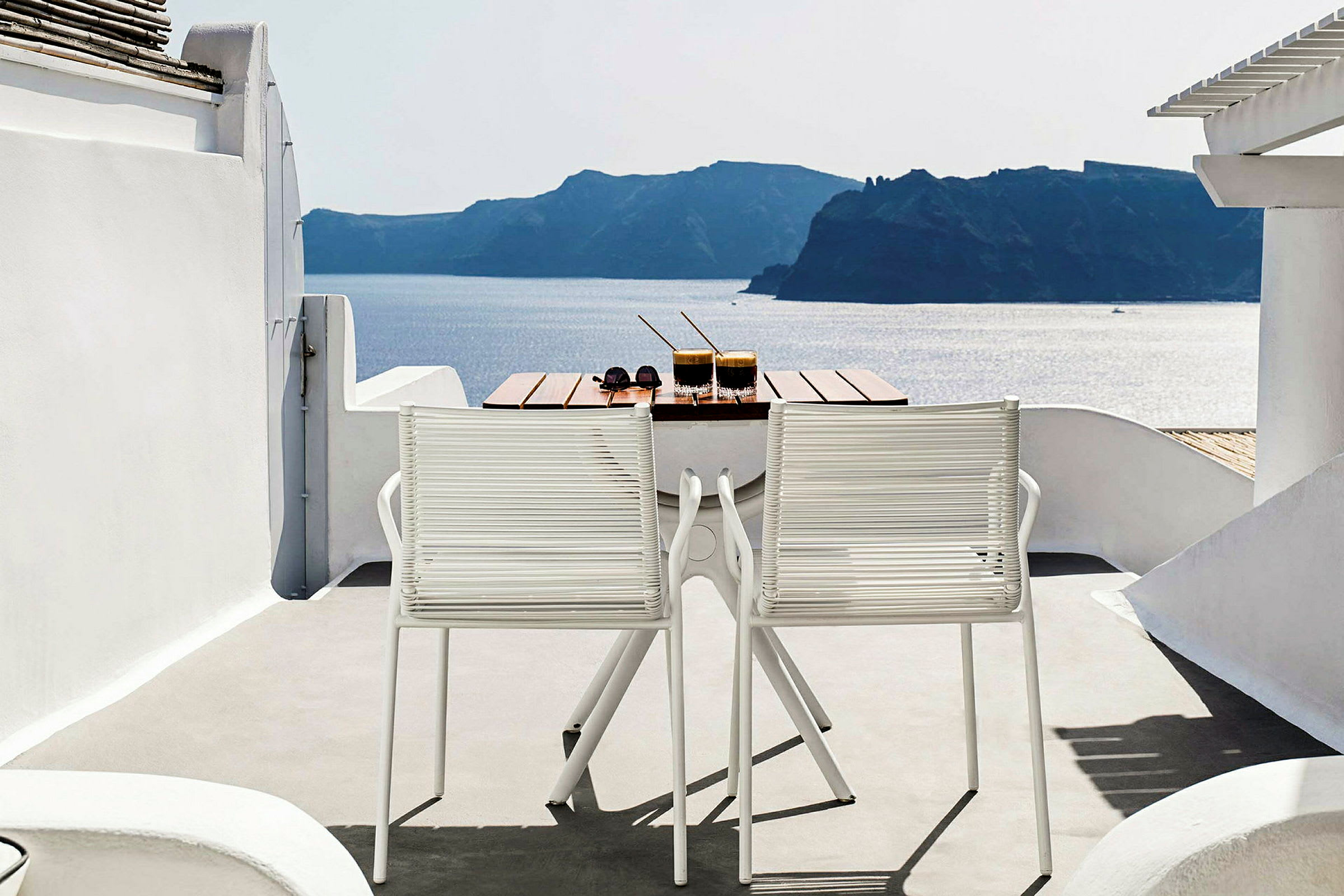 Et bord med to drikkevarer og et par solbriller står på en terrasse med to hvide stole. Udsigten viser havet og landskab i baggrunden. (AI-genereret tekst)