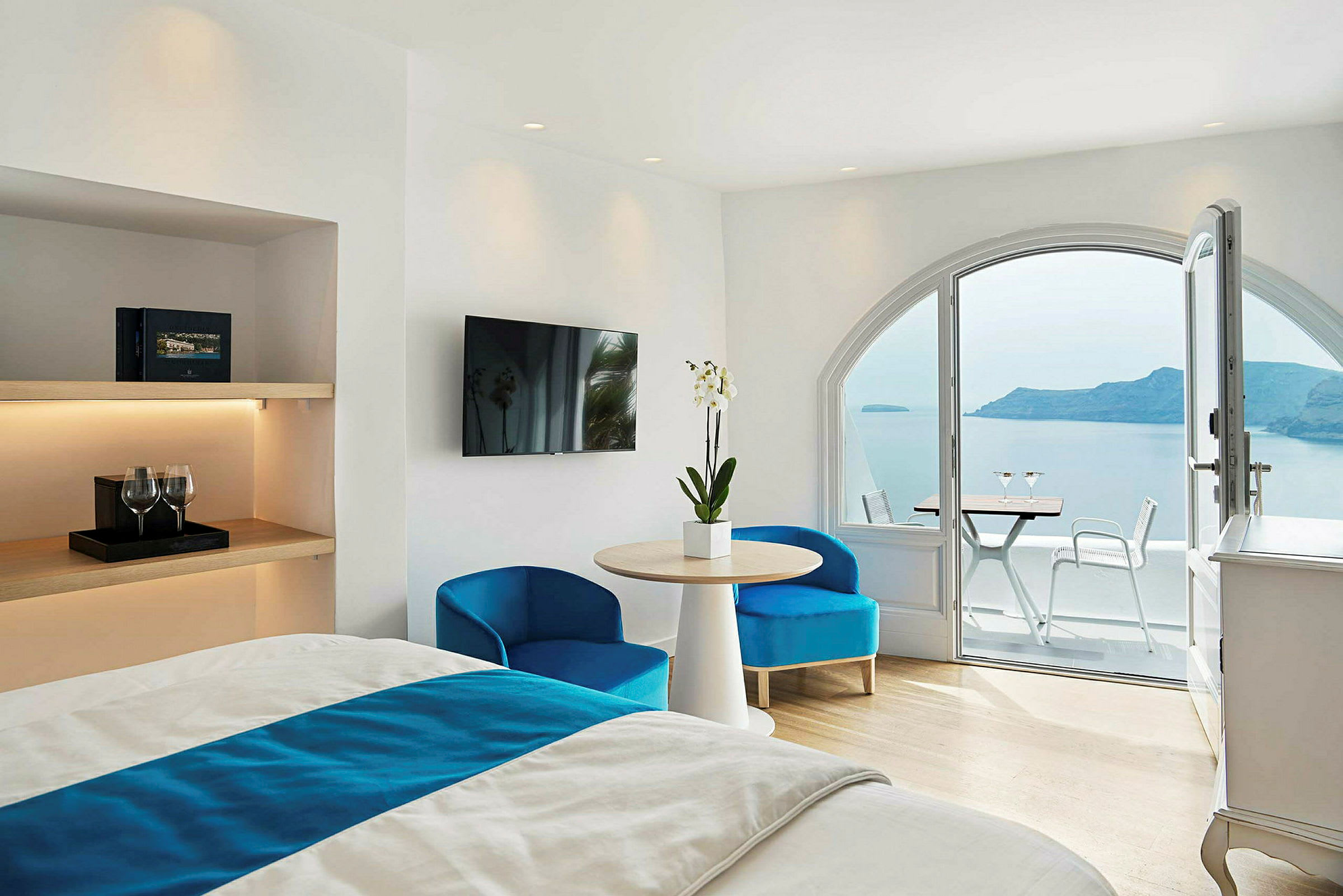 Hotelværelse med lyse hvide vægge, møbleret med en seng, bord og stole, og en balkon med udsigt over havet og en ø. (AI-genereret tekst)