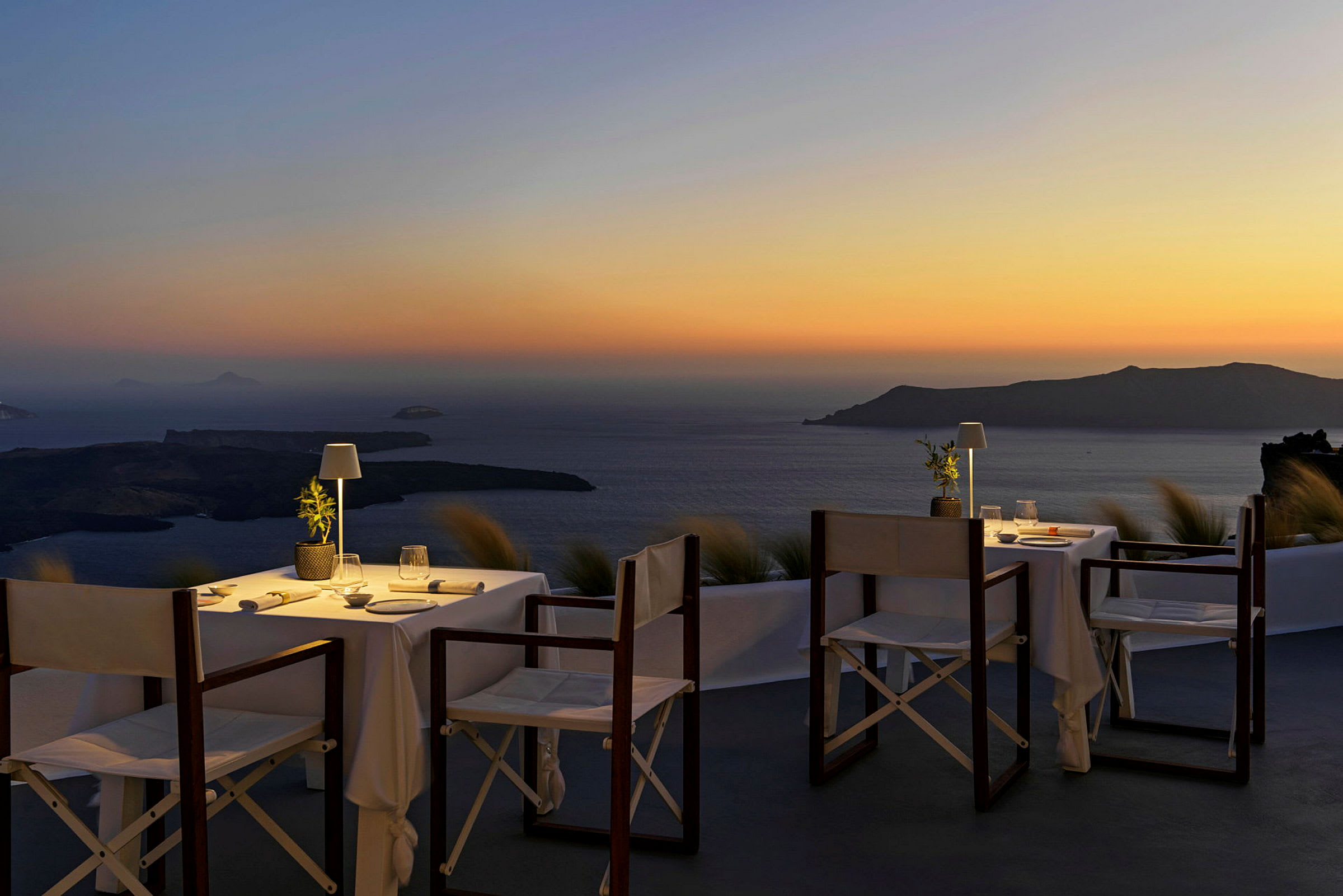 To dækkede borde med stole på en terrasse med udsigt over havet og en ø ved solnedgang. (AI-genereret tekst)