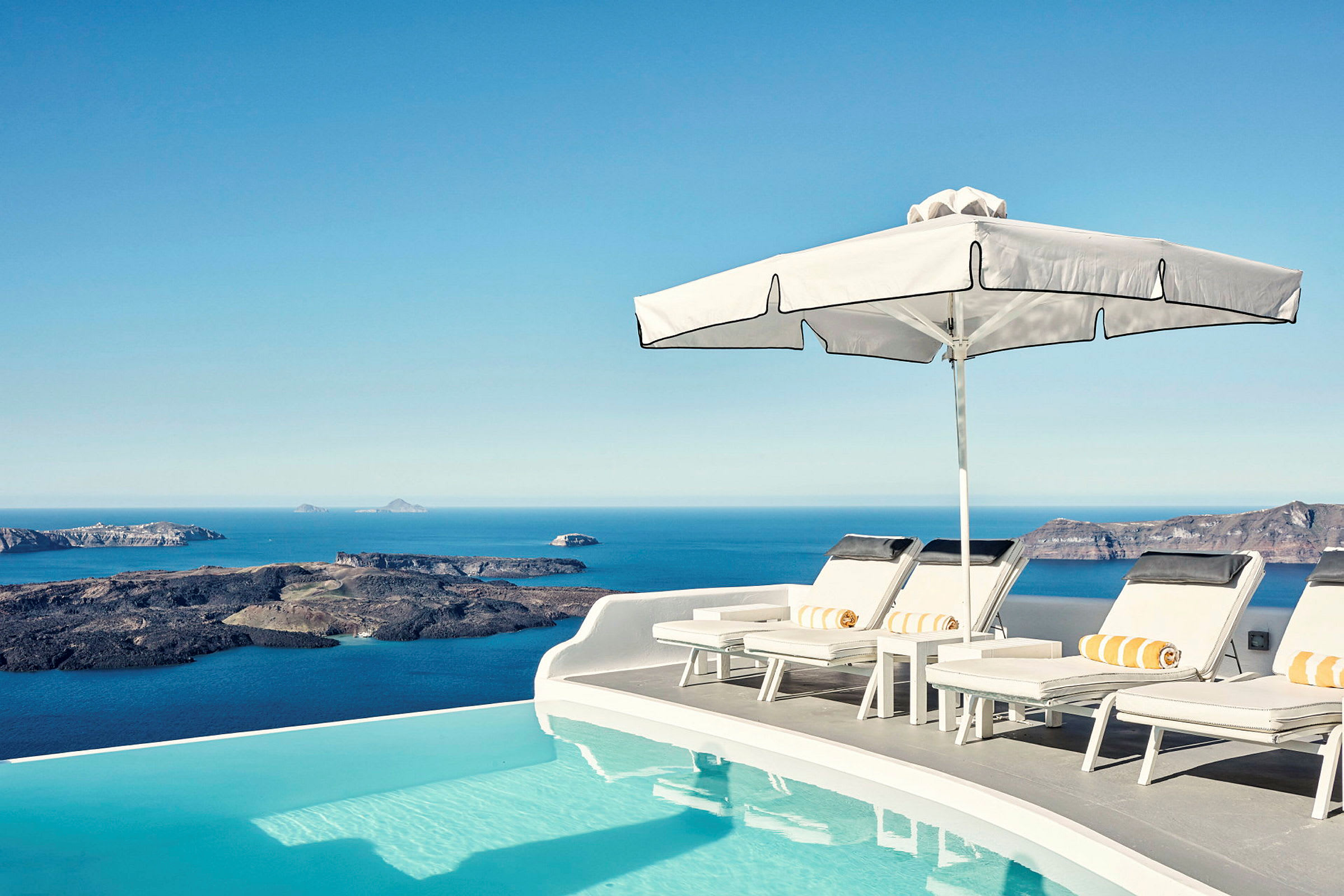 Solterrasse ved en infinitypool med udsigt over havet og klipper i baggrunden. (AI-genereret tekst)