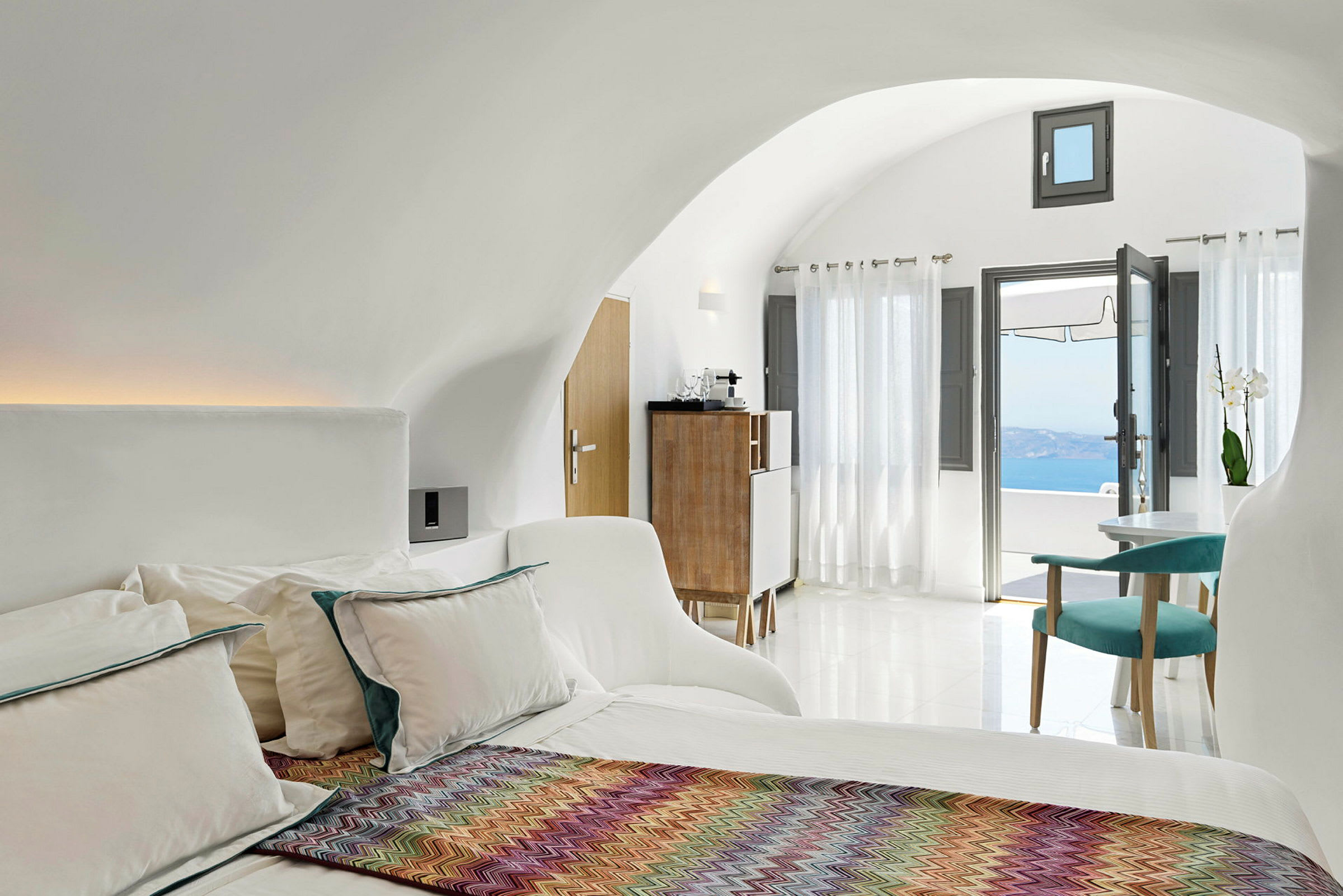 Et lyst og moderne hotelværelse med en seng, farverigt tæppe, siddeområde og store vinduer med udsigt over havet. (AI-genereret tekst)