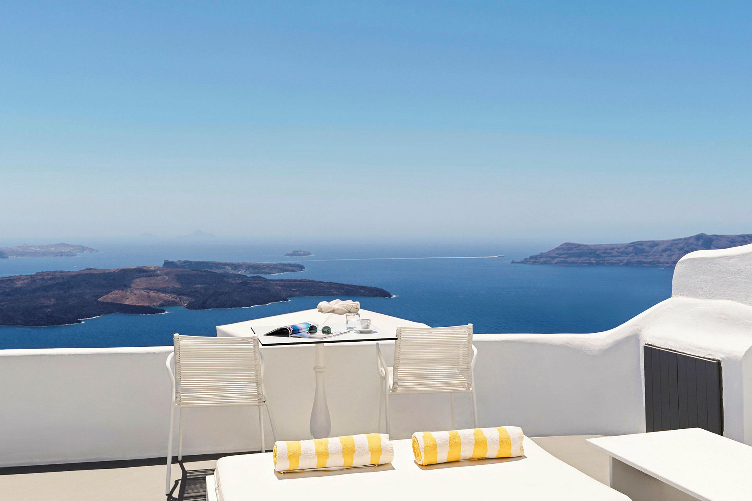 Hvid terrasse med udsigt over havet og øer, med liggestole og et bord med stole. (AI-genereret tekst)