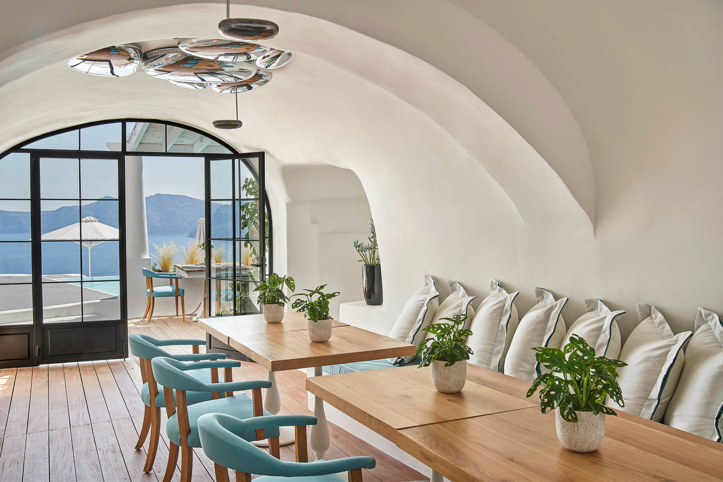 Et elegant interiør med hvælvede lofter, træborde og turkise stole. Store vinduer giver udsigt over en terrasse og havet. (AI-genereret tekst)