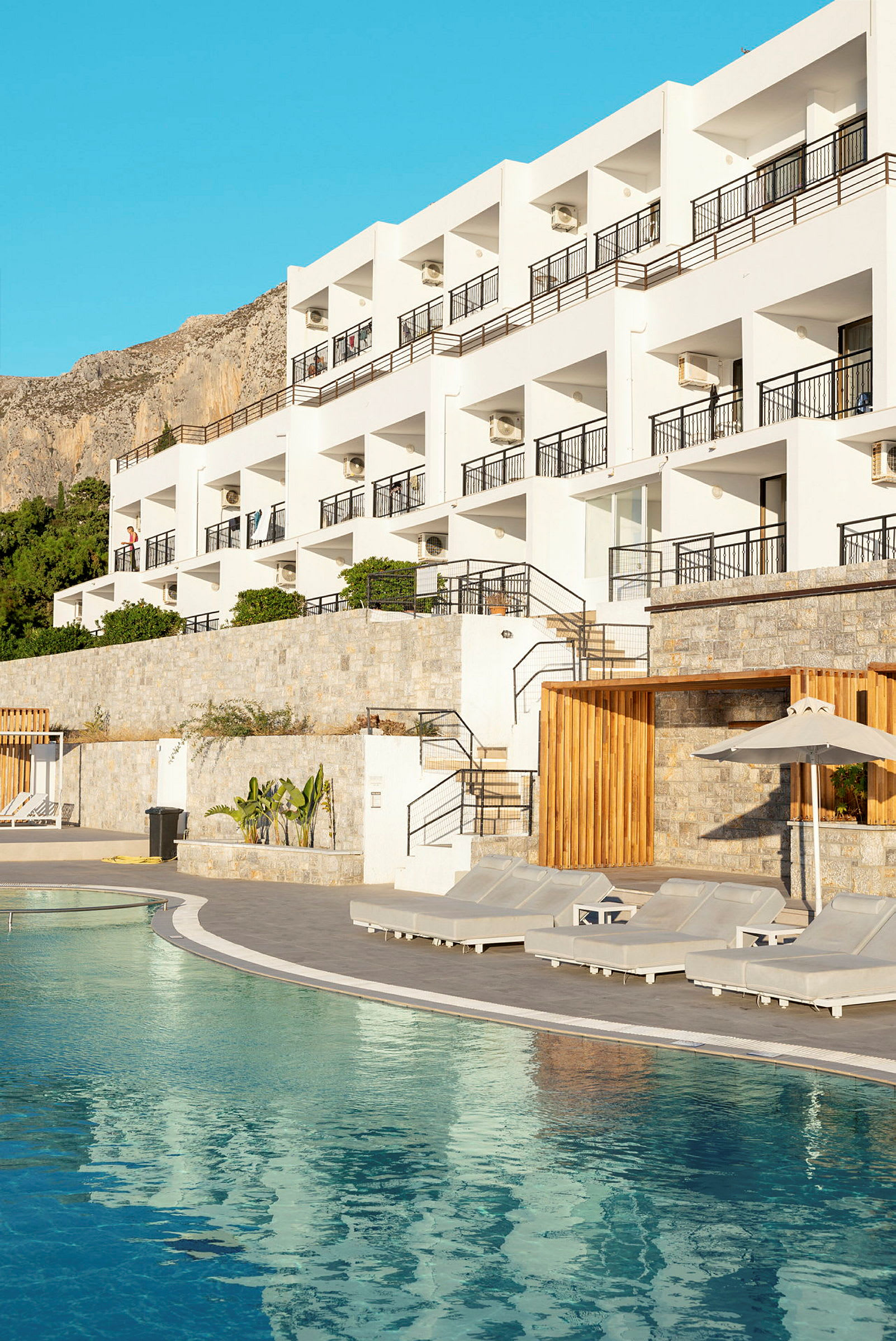 Et hotel ved en pool med liggestole foran. Bygningen har flere altaner og er omgivet af bjerge og en klar blå himmel. (AI-genereret tekst)